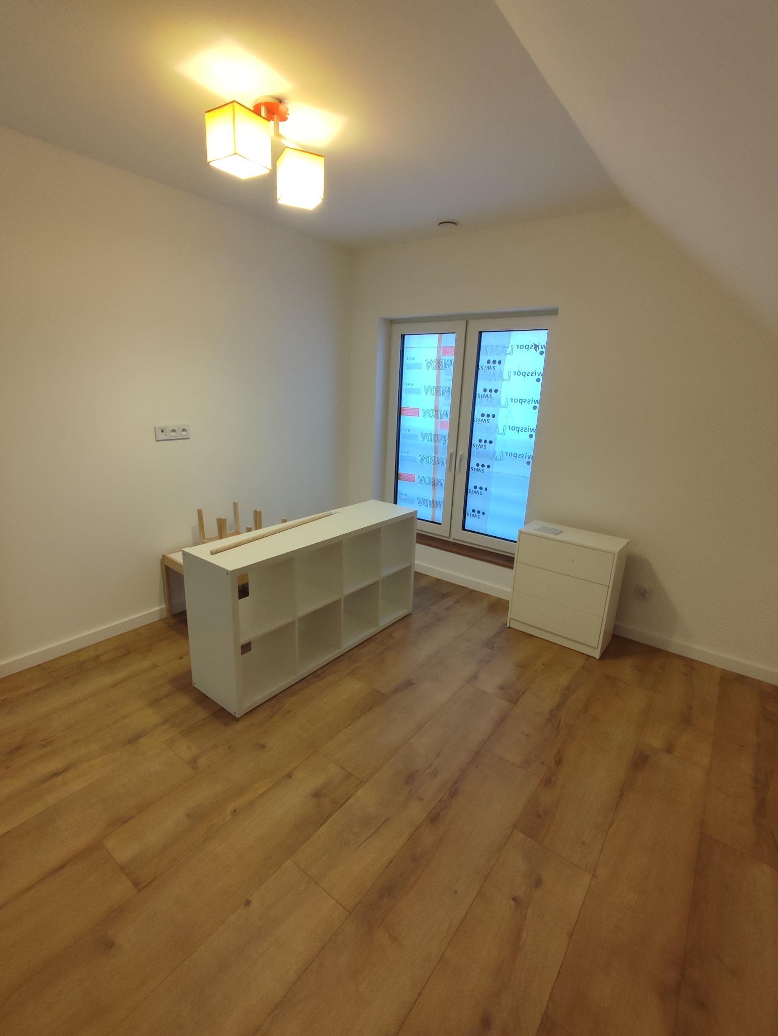 Pusta, jasna sypialnia z drewnianą podłogą, białymi ścianami, oknem zabezpieczonym folią, białym regałem i komodą. Widoczny surowy stan wykończenia wnętrza przed umeblowaniem.