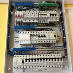 Otwarta skrzynka rozdzielcza z zamontowanymi bezpiecznikami, licznikiem energii i przewodami w kolorach niebieskim, brązowym i zielono-żółtym, umieszczona na żółtej ścianie.