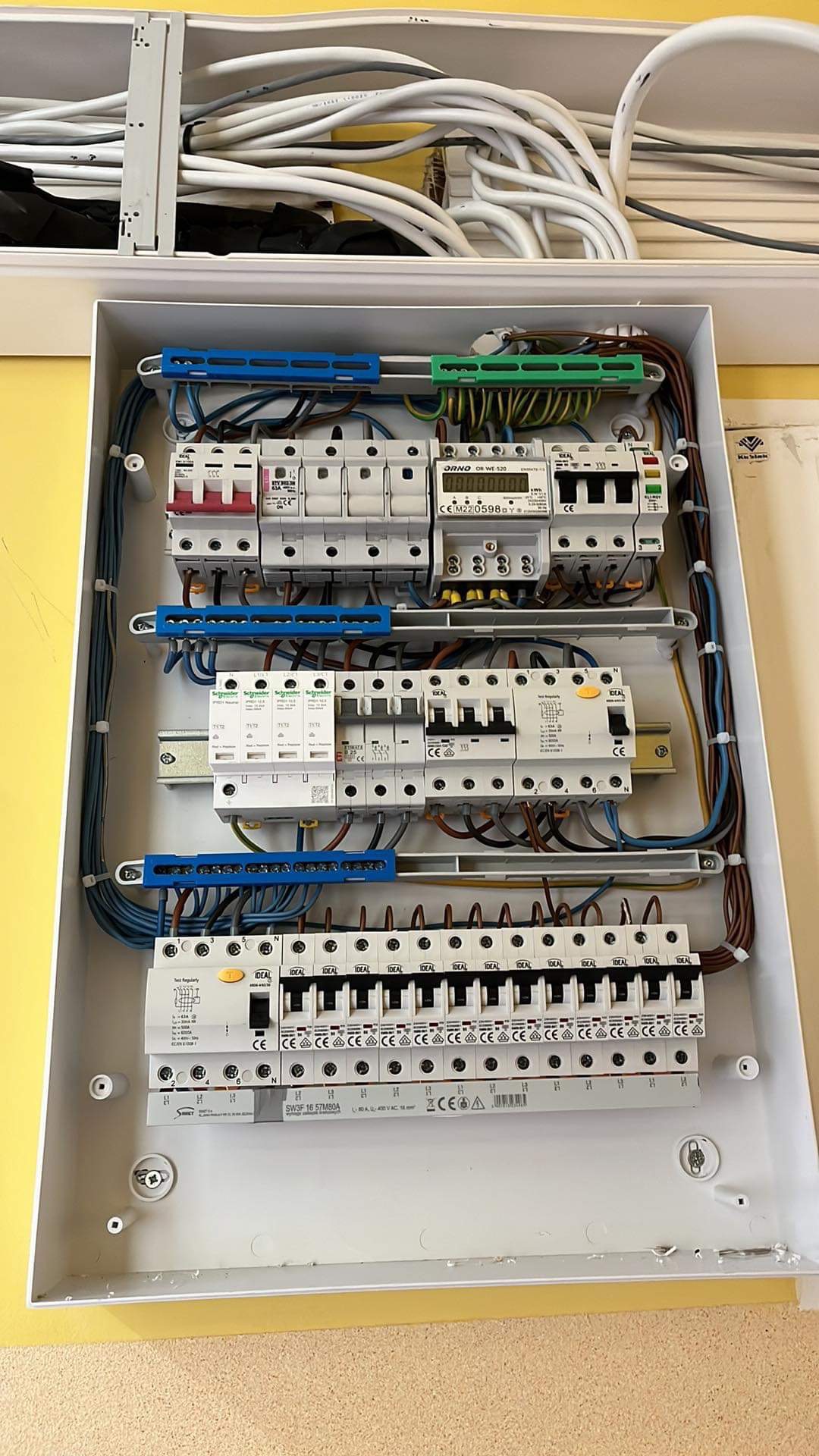 Otwarta skrzynka rozdzielcza z zamontowanymi bezpiecznikami, licznikiem energii i przewodami w kolorach niebieskim, brązowym i zielono-żółtym, umieszczona na żółtej ścianie.