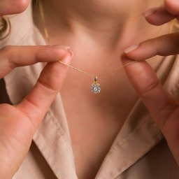 BlackLine Photo Damian Kasperowicz - Delikatny złoty łańcuszek z zawieszką z brylantami, trzymany przez dłonie na tle beżowej koszuli, w subtelnym oświetleniu.