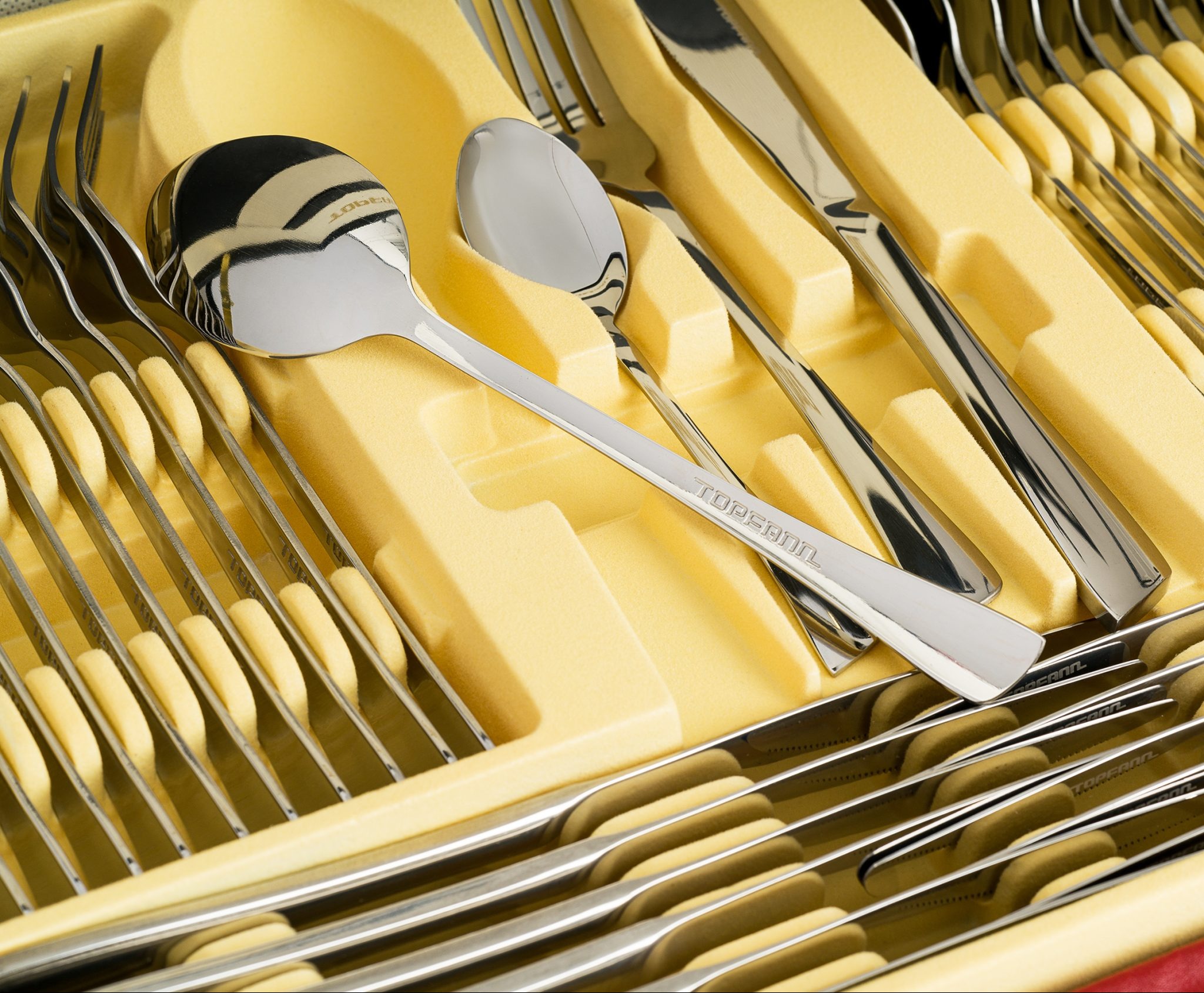 Komplet sztućców marki Topsann w żółtej wkładce, widok z góry, ostre światło podkreśla połysk stali. Precyzyjnie ułożone noże, widelce i łyżki.