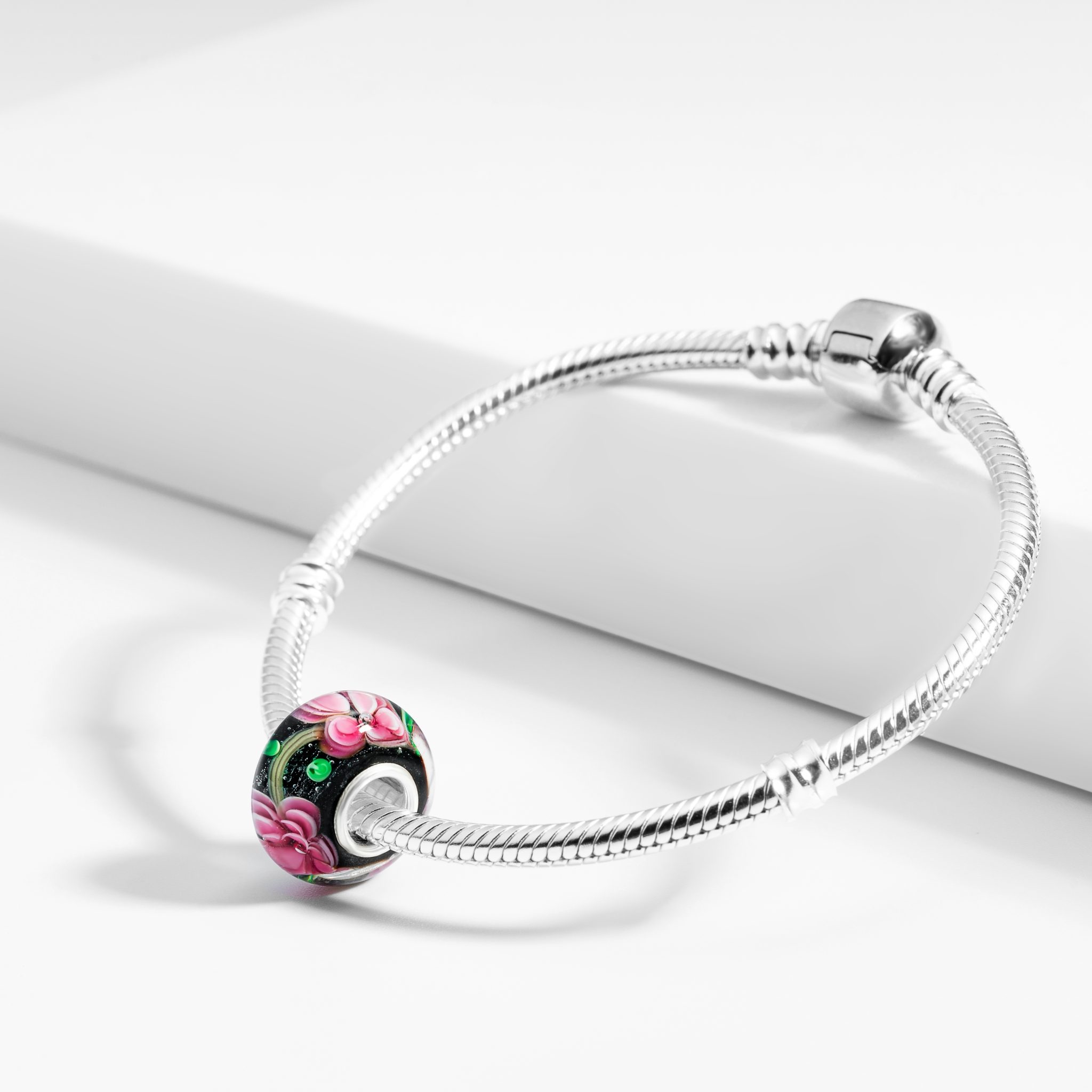 Srebrna bransoletka z ozdobnym, czarnym koralikiem w różowe kwiaty, leżąca na białym tle. Delikatne światło podkreśla detale.