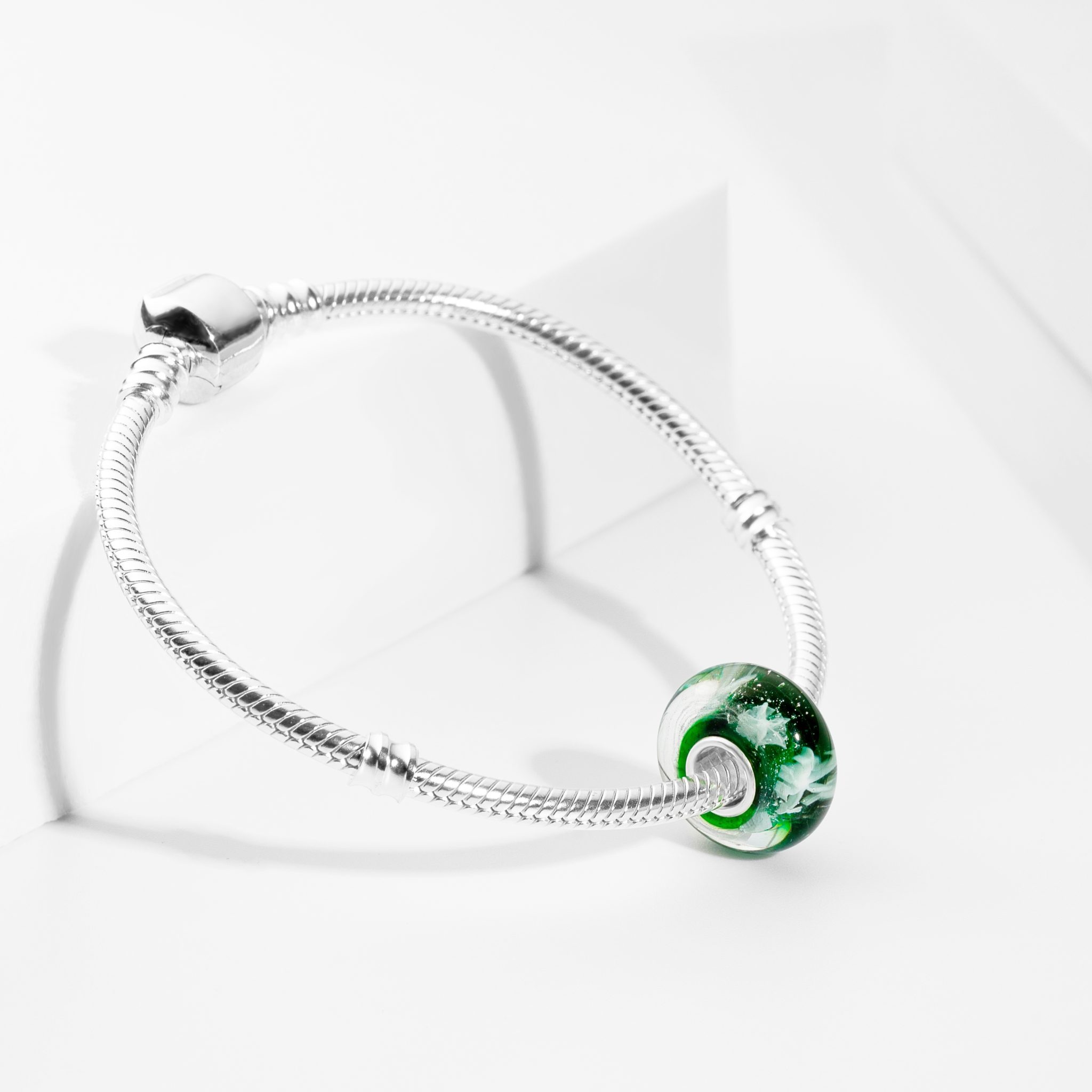 Srebrna bransoletka z zielonym, szklanym charms'em na białym tle. Precyzyjne ujęcie detali, podkreślające fakturę i kolorystykę biżuterii.