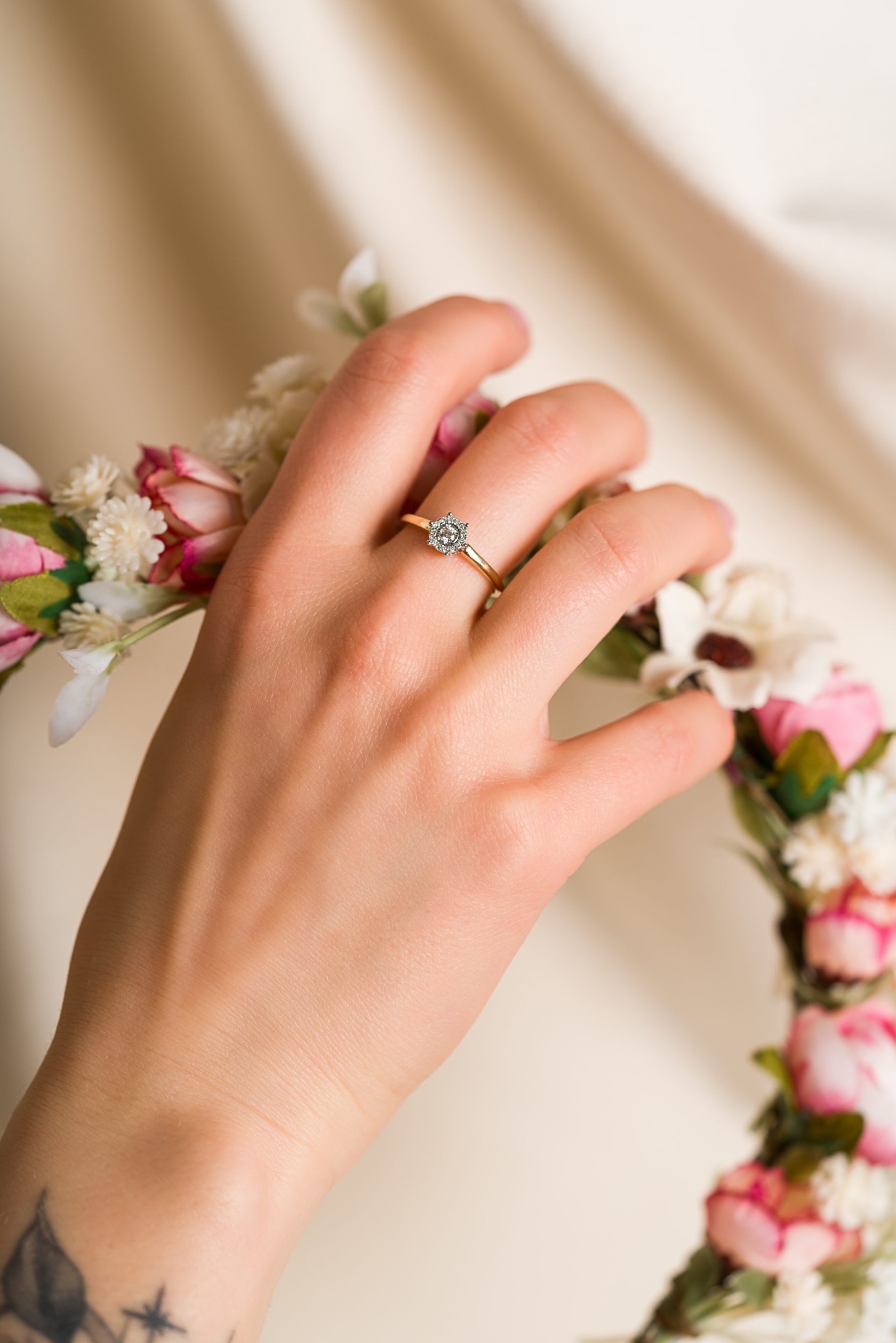 Dłoń z pierścionkiem zaręczynowym na tle kwiatowego wianka w Białymstoku. Delikatne oświetlenie podkreśla detale biżuterii i tatuaż na nadgarstku.