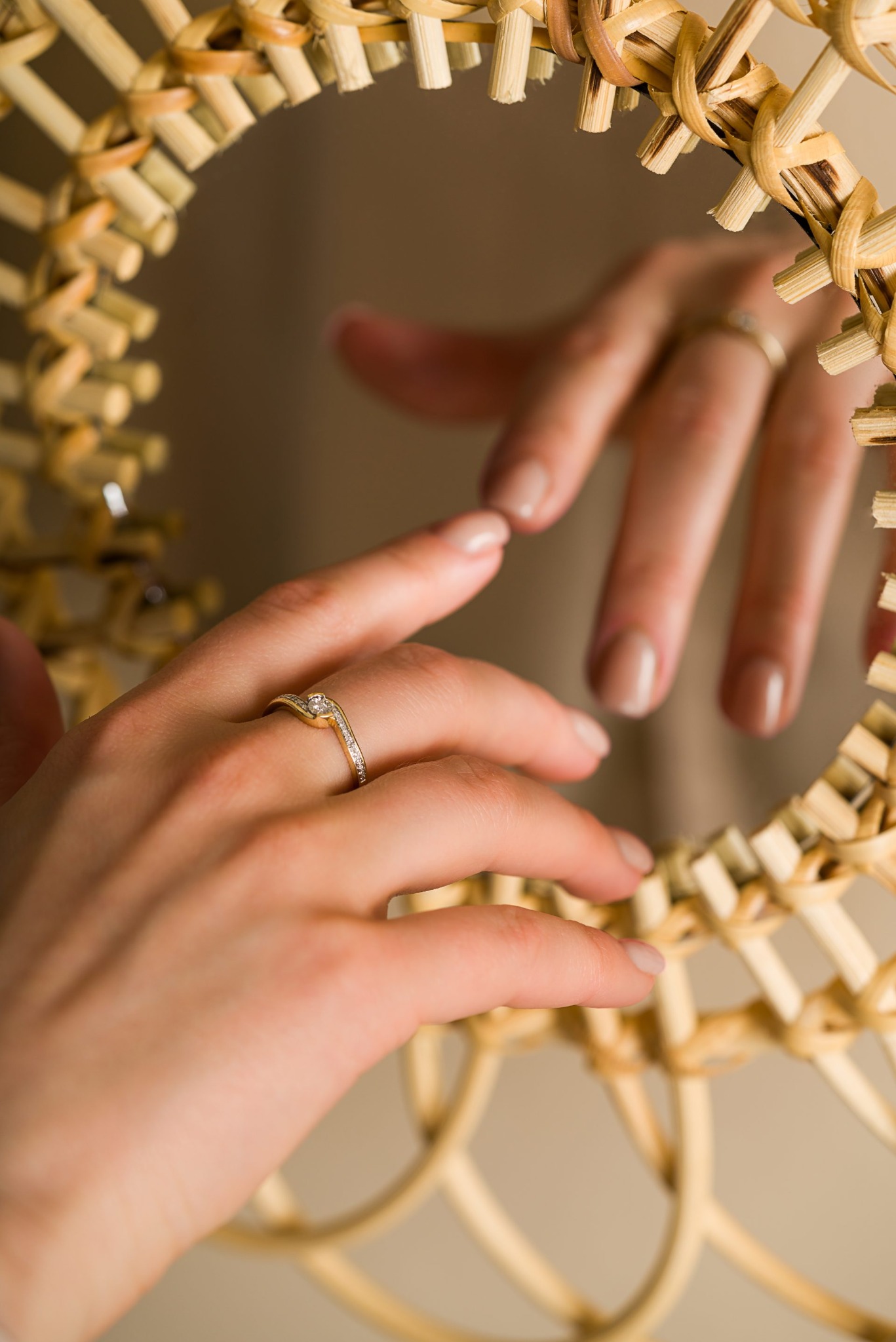 Delikatny pierścionek zaręczynowy na kobiecej dłoni, odbity w wiklinowym lustrze. Subtelny manicure nude. Ciepłe oświetlenie podkreśla blask złota i diamentów.
