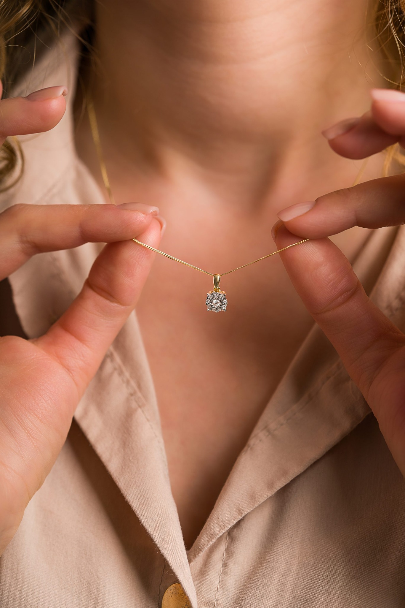 Delikatny złoty łańcuszek z zawieszką z brylantami, trzymany przez dłonie na tle beżowej koszuli, w subtelnym oświetleniu.