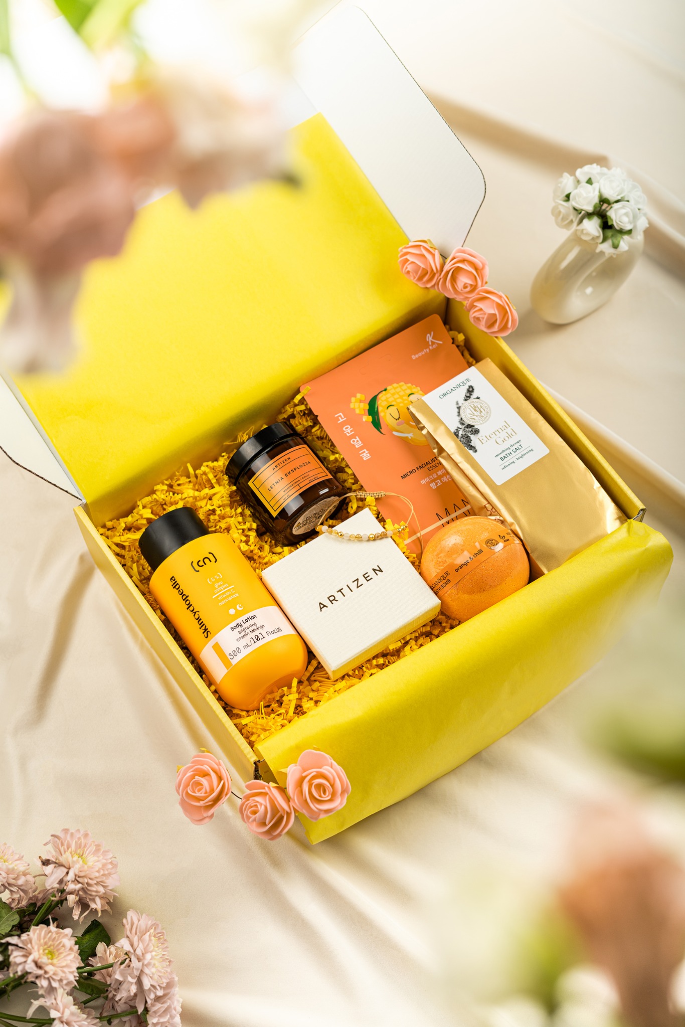 Żółte pudełko prezentowe wypełnione kosmetykami i ozdobami kwiatowymi na kremowym tle, ujęcie z góry, oświetlenie studyjne.