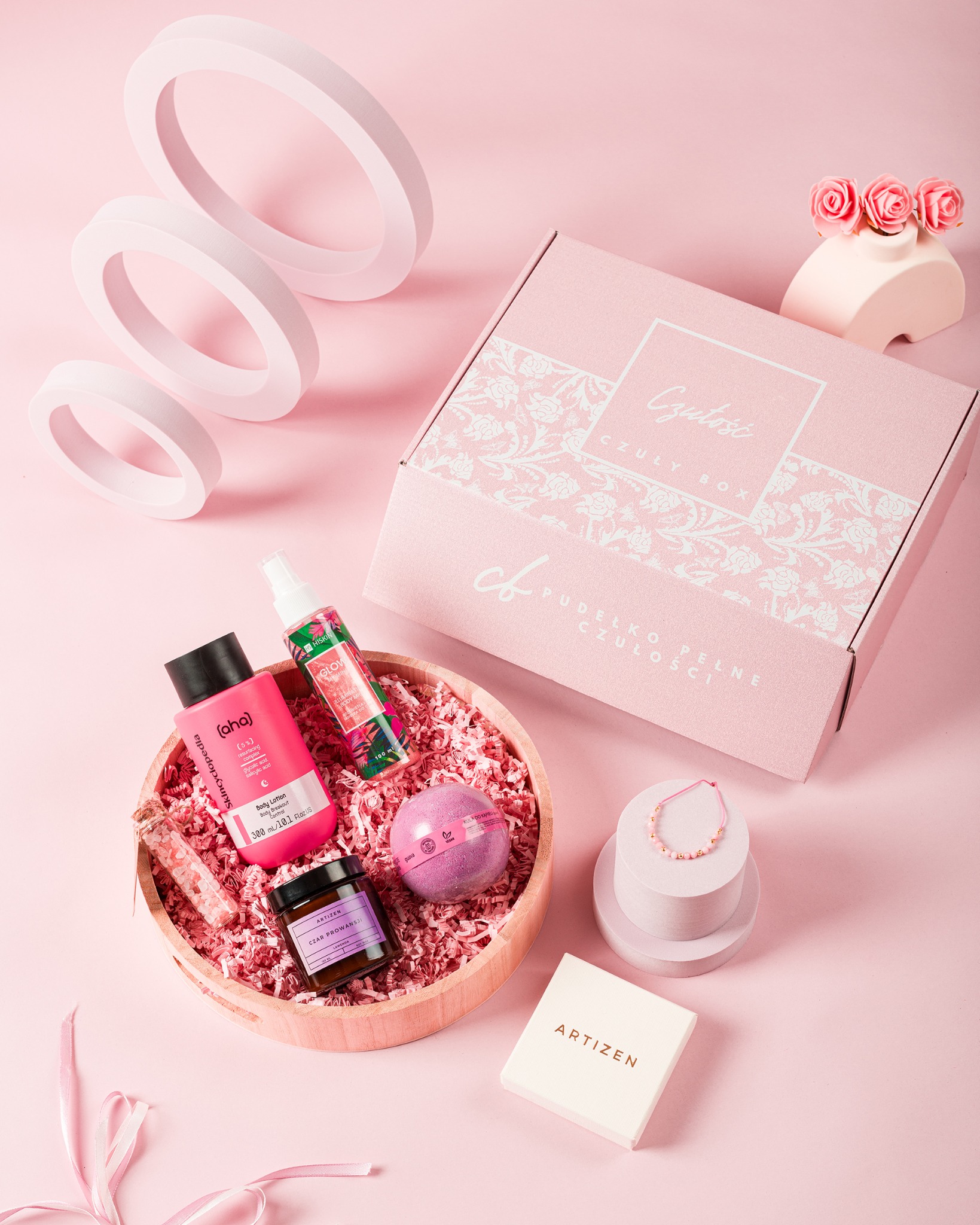 Różowe tło z pudełkiem 'Czułość', kosmetykami i biżuterią w drewnianej misie. Dekoracyjne okręgi i róże w tle. Prezentacja zestawu upominkowego.