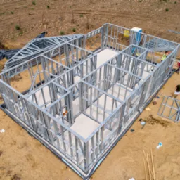 Widok z góry na szkielet stalowy budowanego domu jednorodzinnego z widocznymi pracownikami w kamizelkach odblaskowych, przygotowanymi elementami konstrukcji dachu i przenośną toaletą.