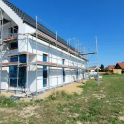 Nowoczesny budynek jednorodzinny w trakcie budowy, z białymi ścianami, ciemną dachówką i rusztowaniami, okna zabezpieczone folią, widok z zewnątrz w słoneczny dzień.