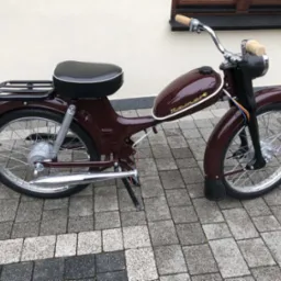 Odnawiany motorower Komar w kolorze bordowym z chromowanymi elementami, zaparkowany na kostce brukowej przed budynkiem z oknem.