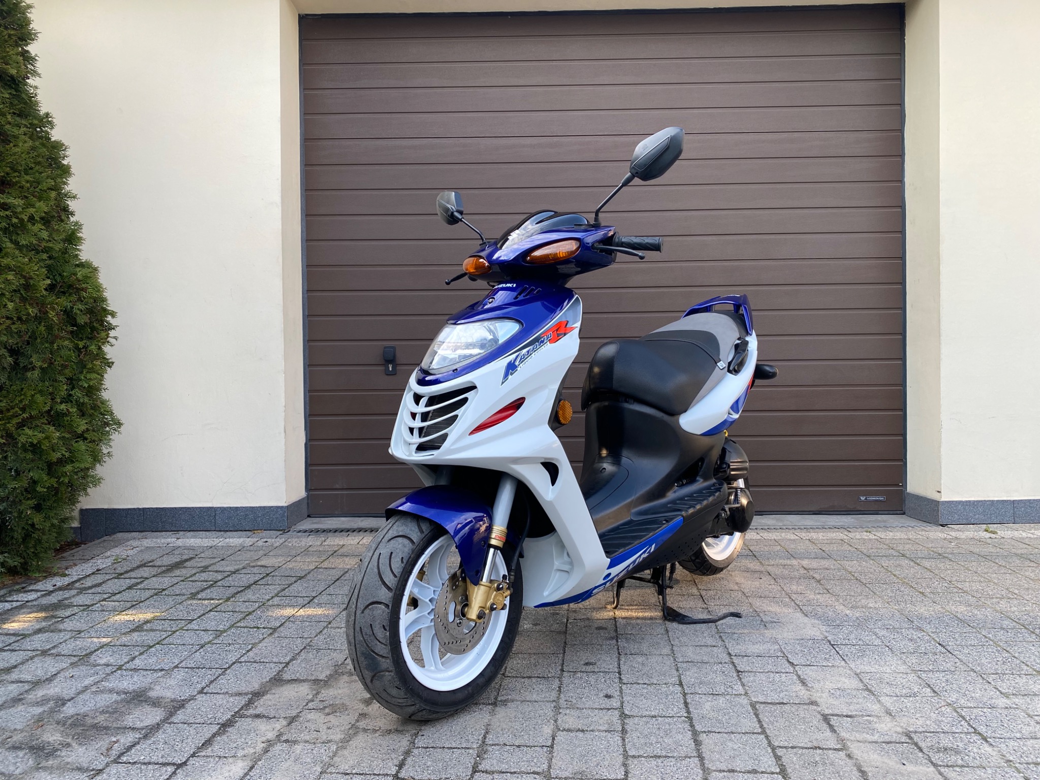 Biało-niebieski skuter Suzuki Katana R na kostce brukowej przed brązową bramą garażową i tujami.