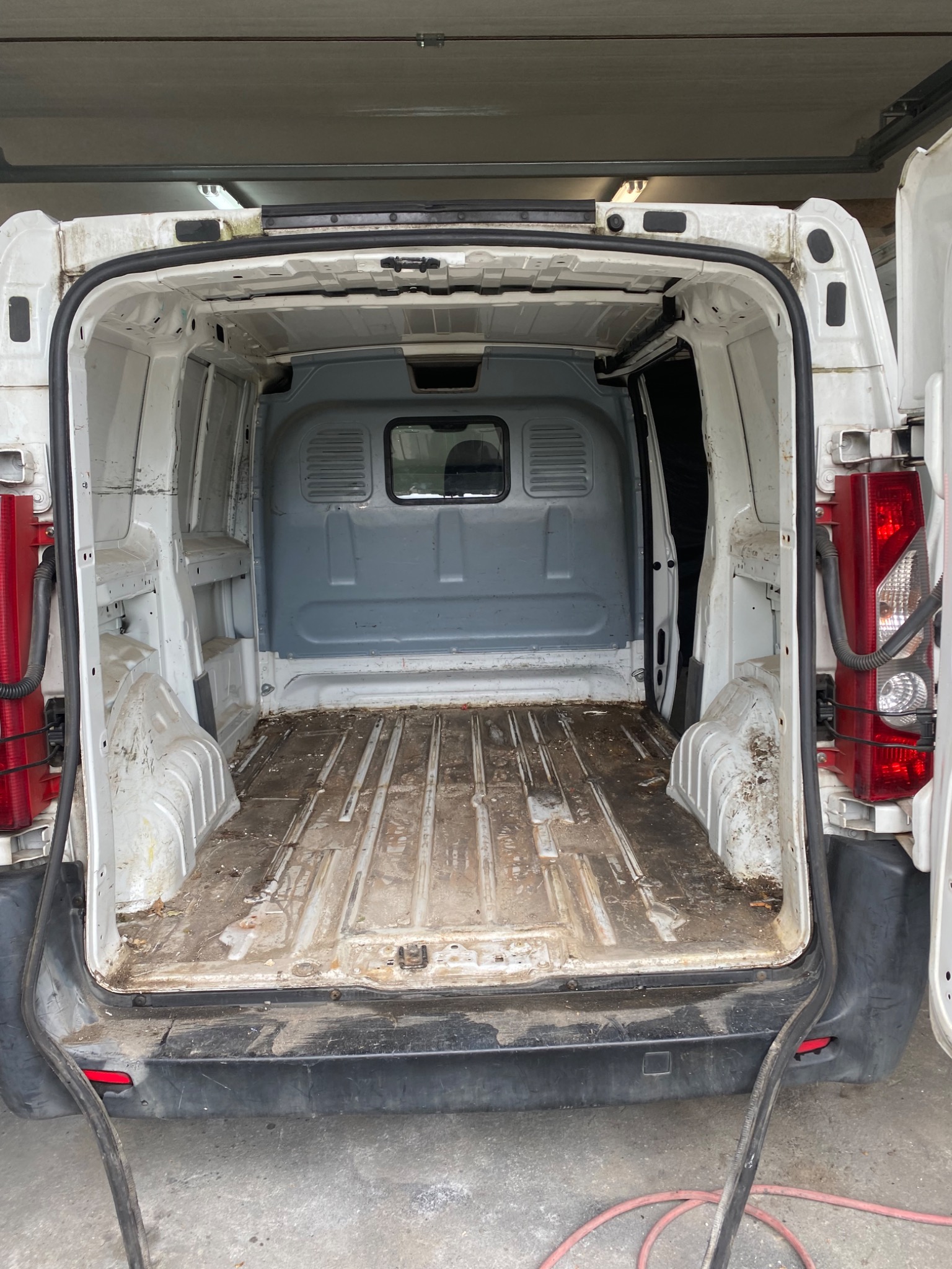 Wnętrze brudnego, białego furgonetki z otwartymi drzwiami, widoczne ślady użytkowania na podłodze i ścianach, gumowa uszczelka zwisa z tylnego zderzaka.