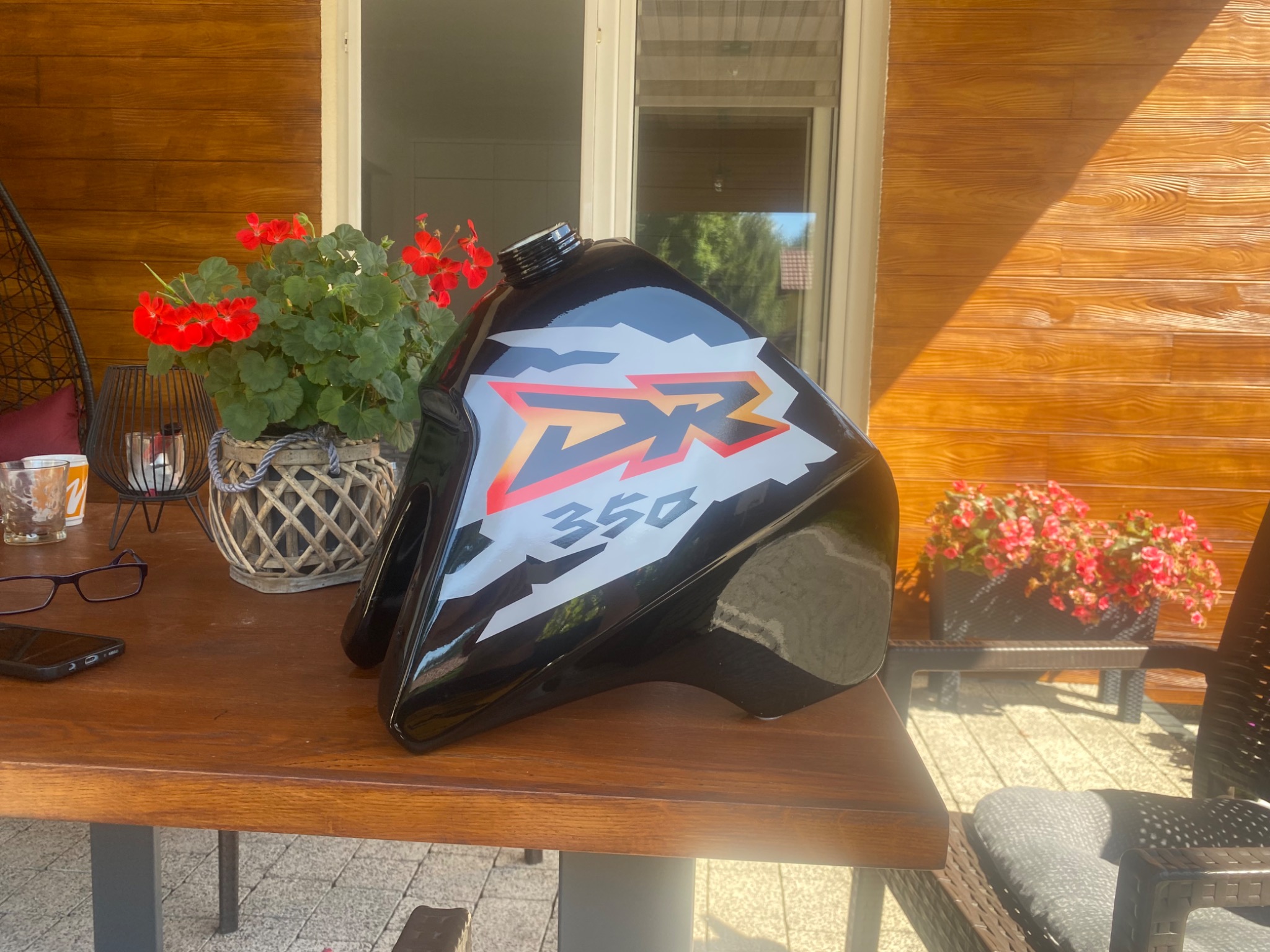 Czarny, błyszczący bak motocyklowy DR 350 po renowacji, leżący na drewnianym stole na tle drewnianej elewacji domu i kwiatów w doniczkach.