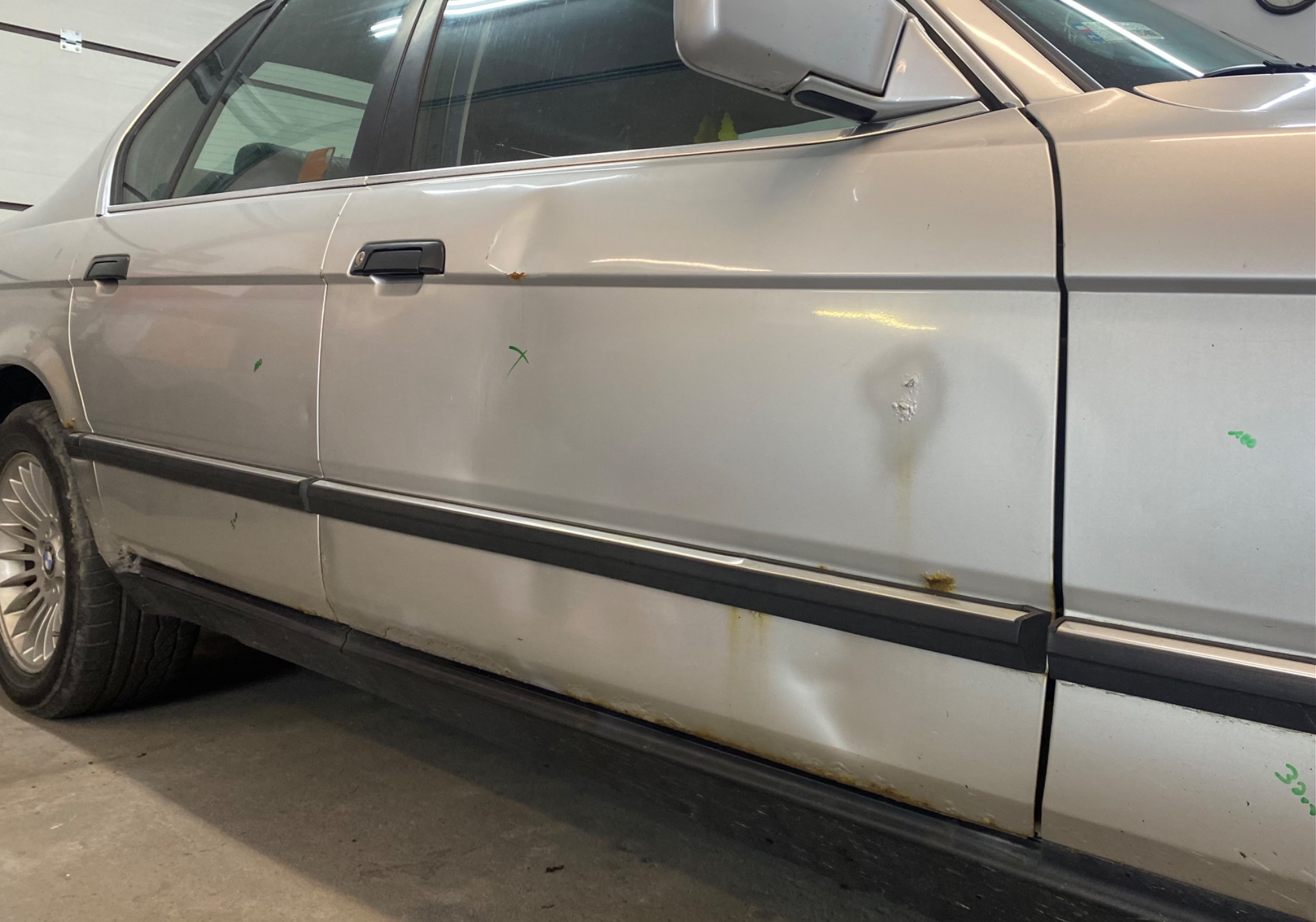 Srebrny samochód z widocznymi uszkodzeniami karoserii, wgnieceniami i rdzawymi plamami, zaznaczone zielonymi znakami w warsztacie.