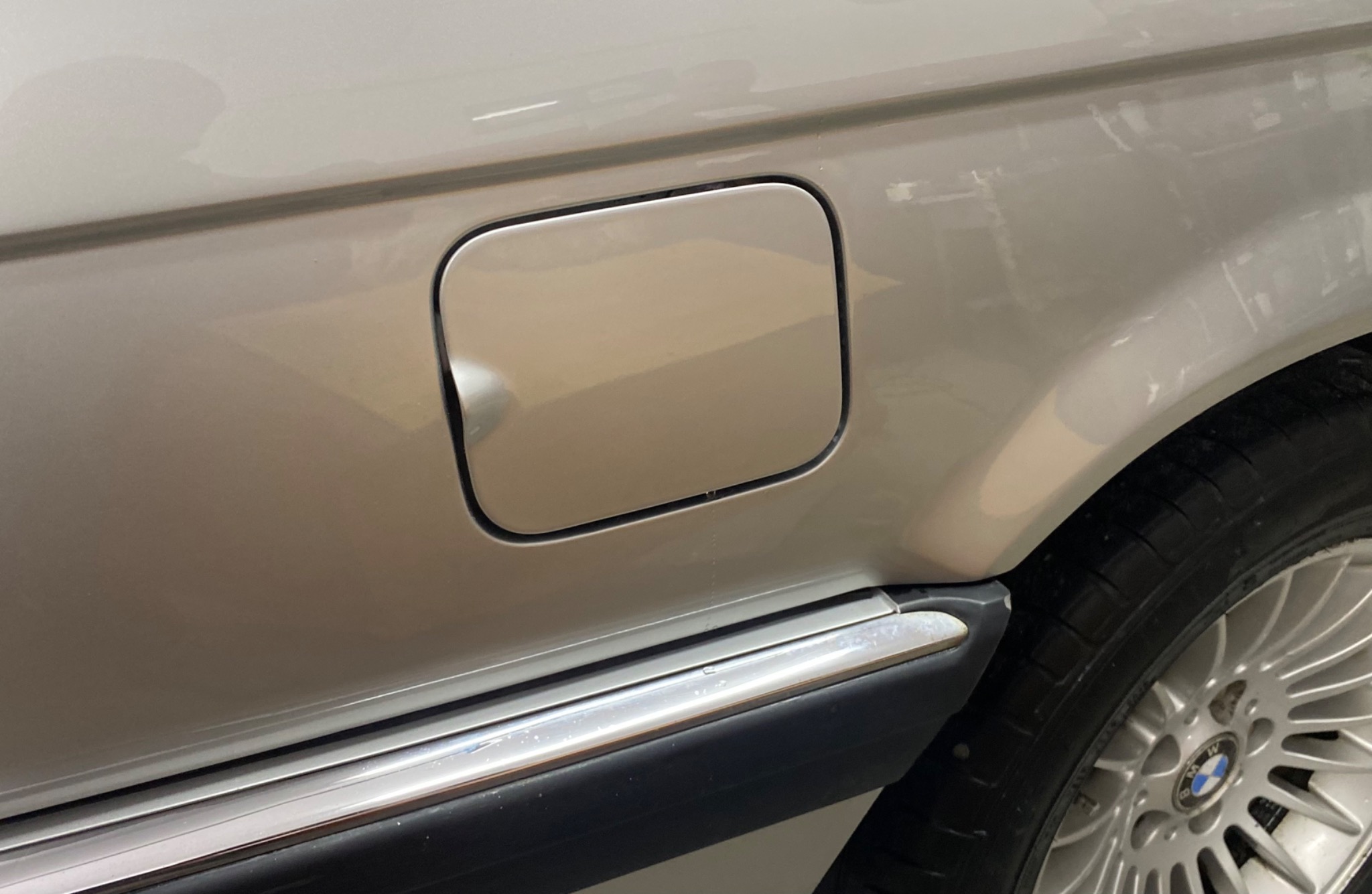 Fragment karoserii srebrnego samochodu z widocznym wlewem paliwa i fragmentem opony z felgą.