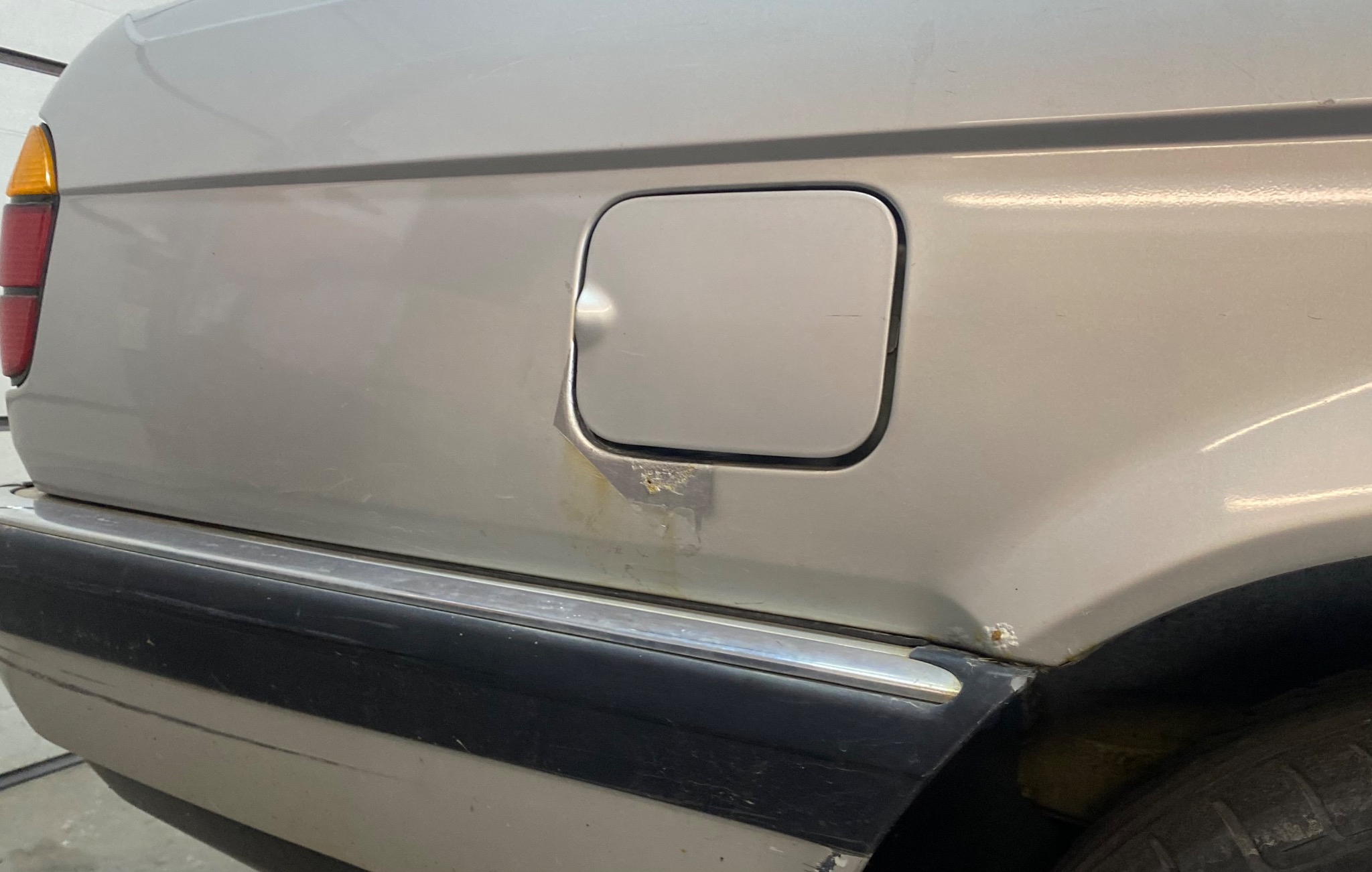 Fragment srebrnego samochodu z widoczną korozją wokół klapki wlewu paliwa i wgnieceniem na klapce, czarny plastikowy zderzak z chromowaną listwą, widoczny fragment opony.