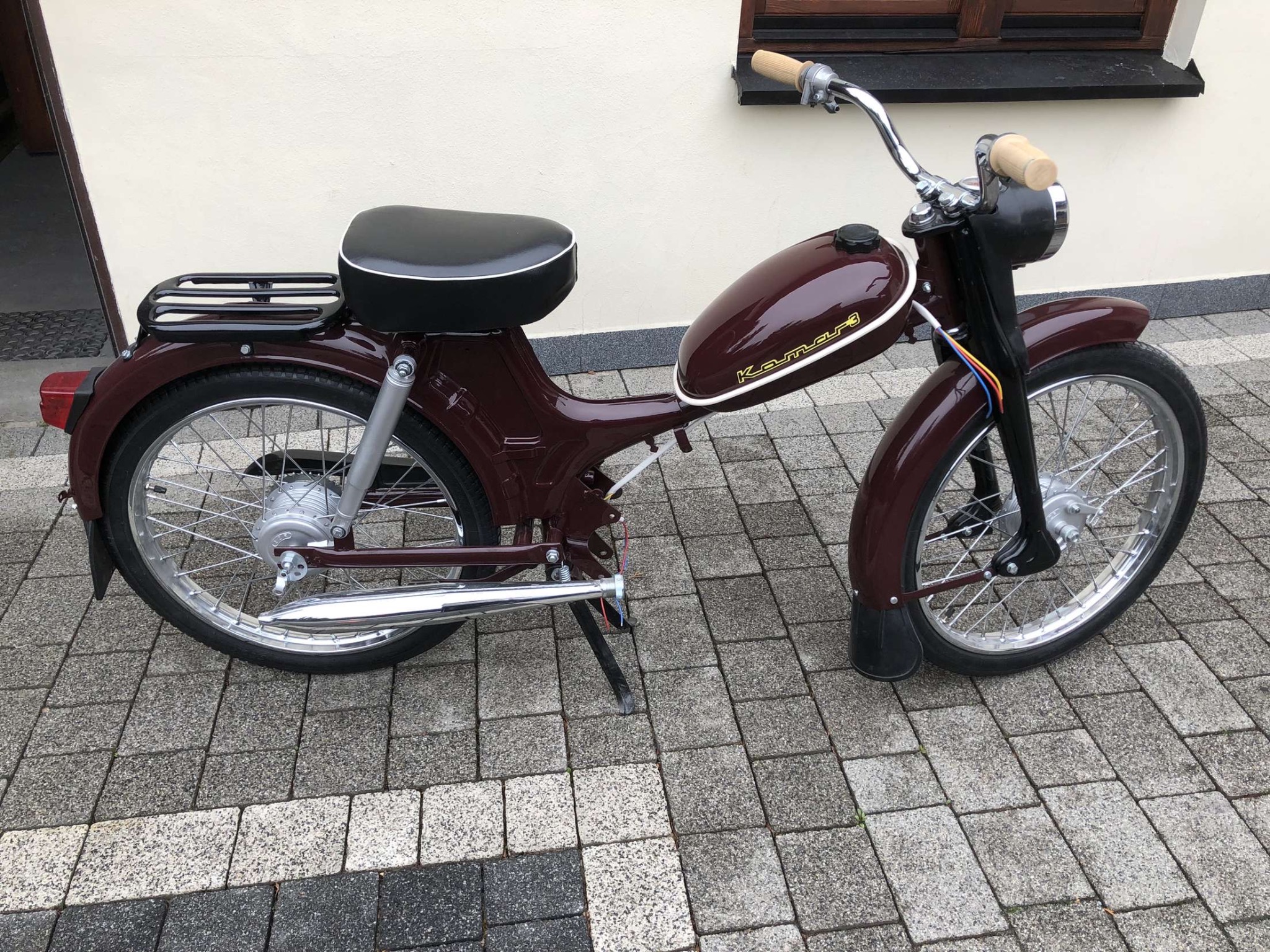 Odnawiany motorower Komar w kolorze bordowym z chromowanymi elementami, zaparkowany na kostce brukowej przed budynkiem z oknem.