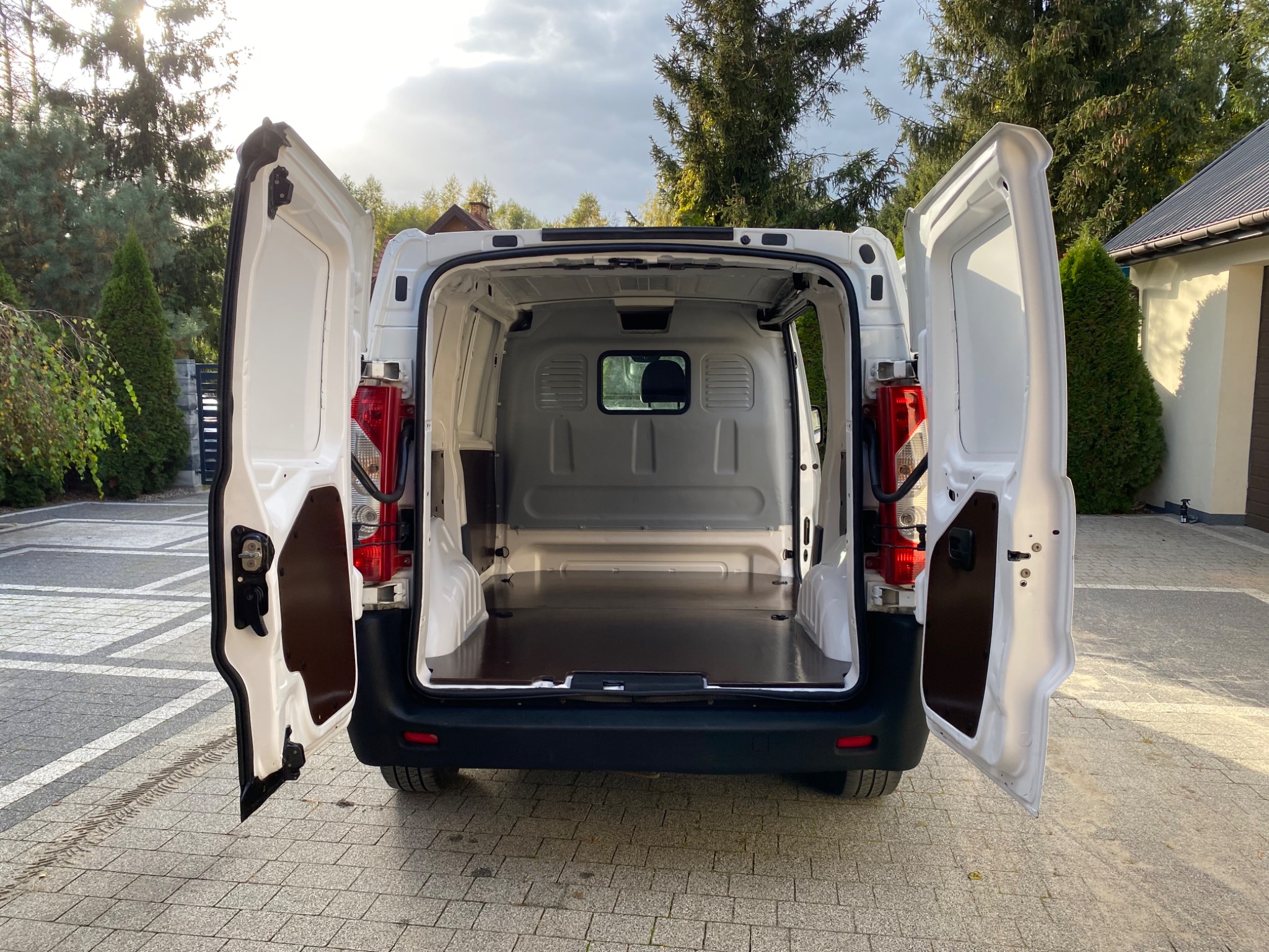 Wnętrze przestrzeni ładunkowej białego furgonu z otwartymi tylnymi drzwiami, widok od tyłu na wyłożoną sklejką podłogę, na zewnątrz kostka brukowa i zieleń
