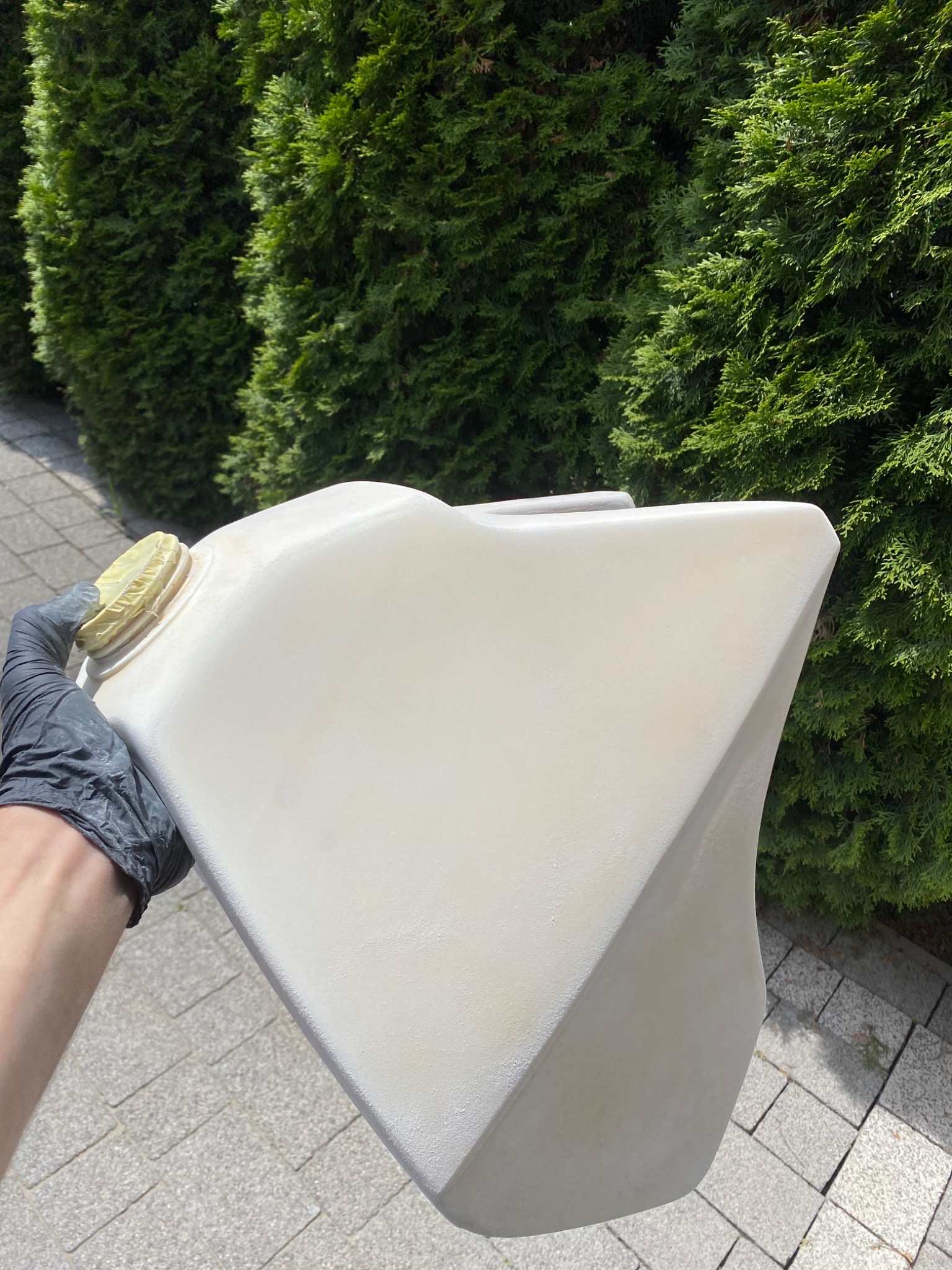 Biały, plastikowy zbiornik paliwa motocykla, trzymany w czarnej rękawicy ochronnej na tle brukowanej ścieżki i żywopłotu z tui.