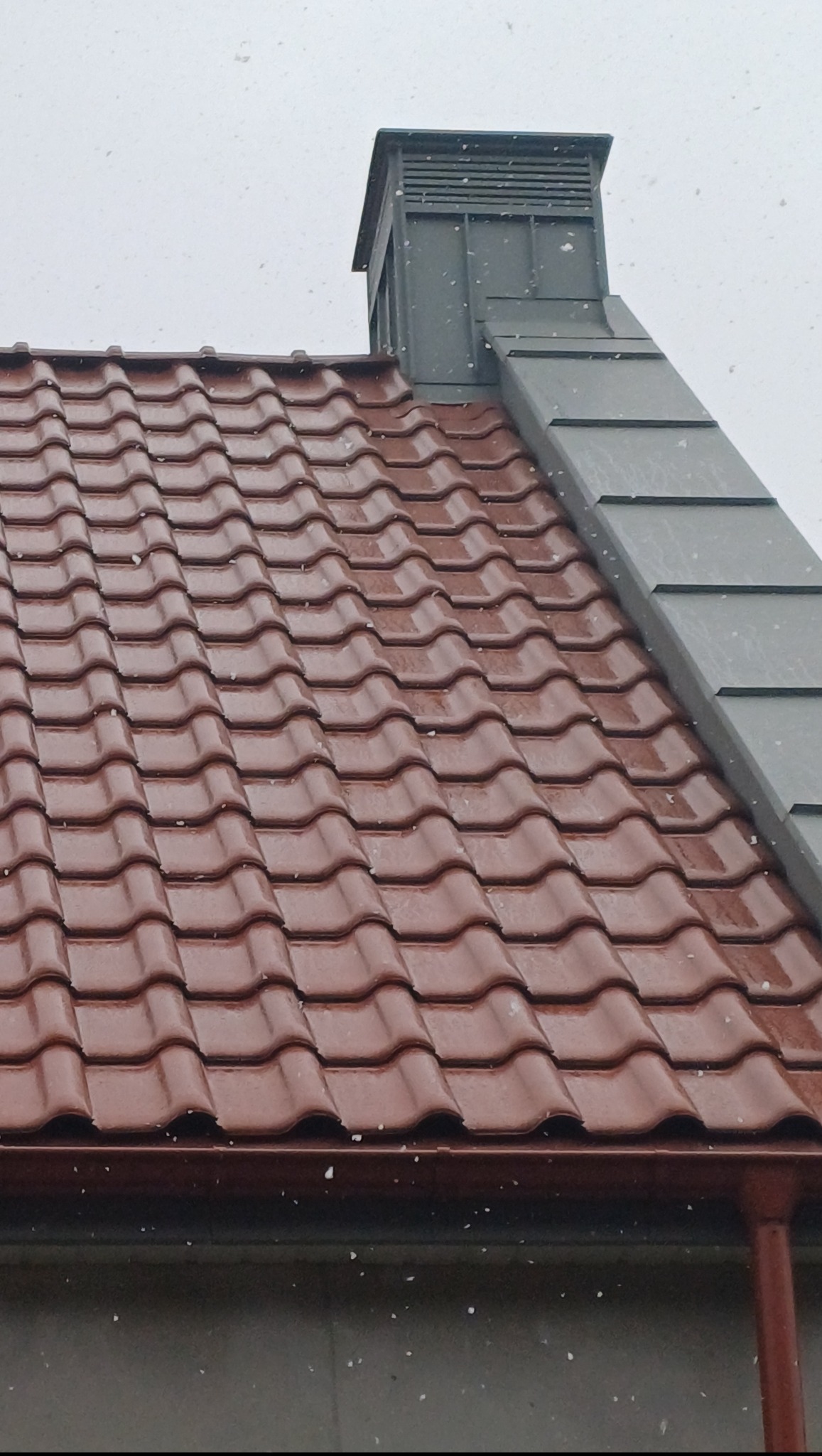 Czerwona dachówka ceramiczna na dachu domu, z widocznym kominem obłożonym blachą w kolorze szarym. Na dachu i w powietrzu widoczne opady śniegu.