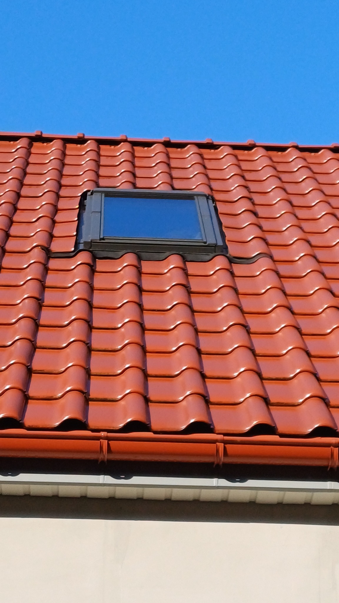 Czerwona dachówka ceramiczna z oknem dachowym i rynną w słoneczny dzień. Widok z dołu na fragment dachu i błękitne niebo.