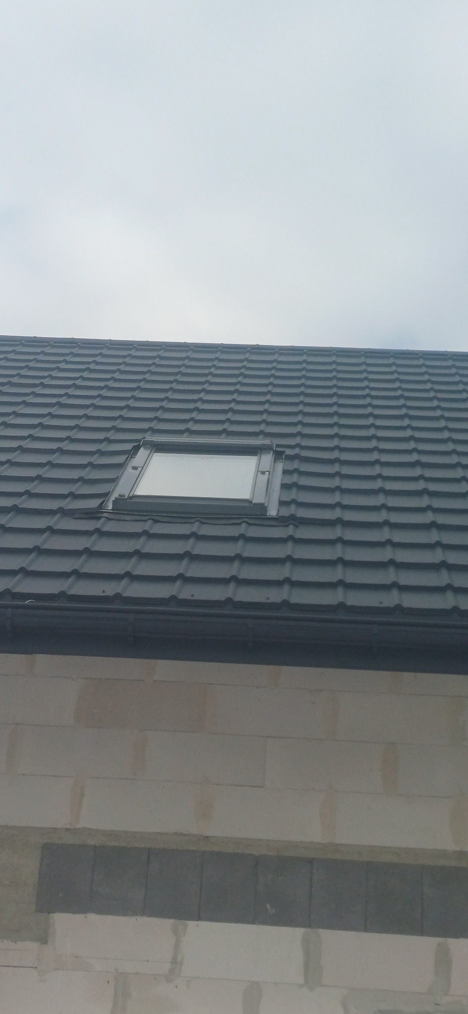 Szary dach z dachówki z oknem dachowym, widok z dołu na tle pochmurnego nieba. Widoczny fragment ściany budynku z szarego bloczka.