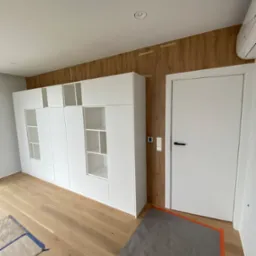 Biała, modułowa zabudowa meblowa z otwartymi p&oacute;łkami i szafkami, dopasowana do ściany z drewnopodobnym panelem, widoczny fragment podłogi zabezpieczony folią malarską.