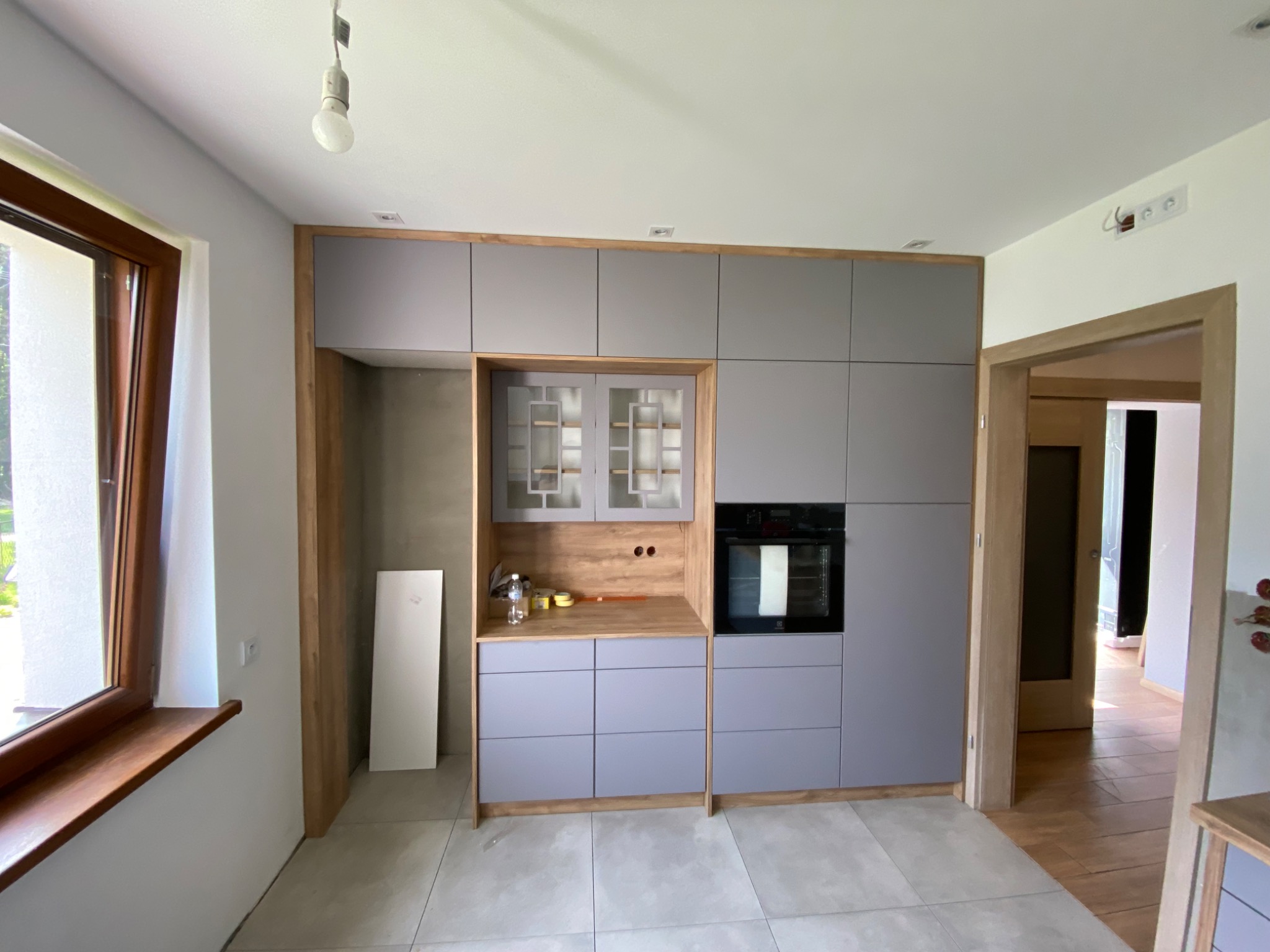 Szare szafki kuchenne z drewnianymi akcentami, częściowo zamontowane, z widoczną wnęką na lodówkę i piekarnikiem, na tle jasnych ścian i podłogi z płytek.