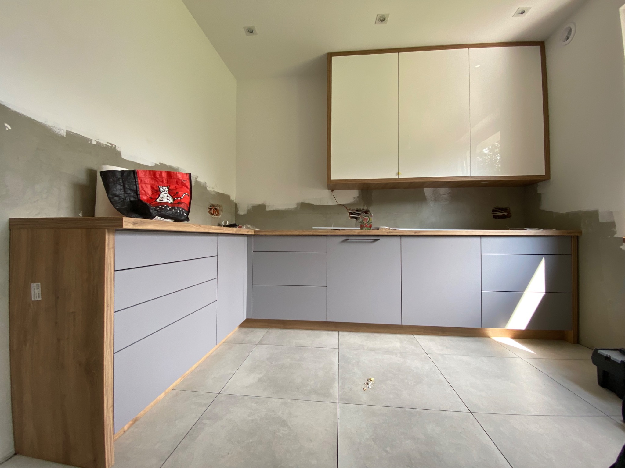 Montaż szarej kuchni na wymiar z drewnianymi blatami i górnymi szafkami w trakcie wykończenia, widoczna torba na blacie i niedomalowane ściany.