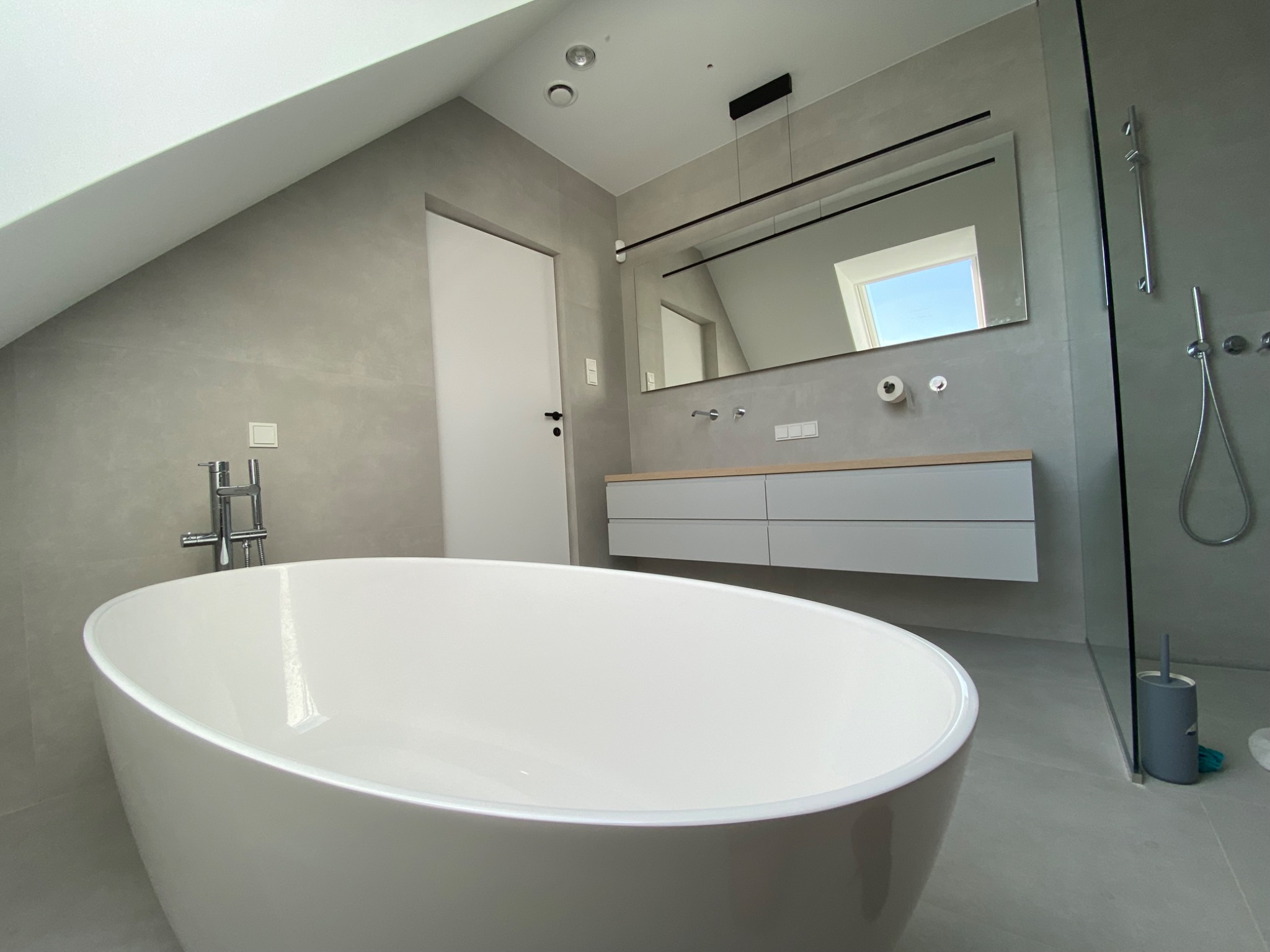 Nowoczesna łazienka na poddaszu z wolnostojącą białą wanną, szarą zabudową umywalkową i minimalistycznym oświetleniem.