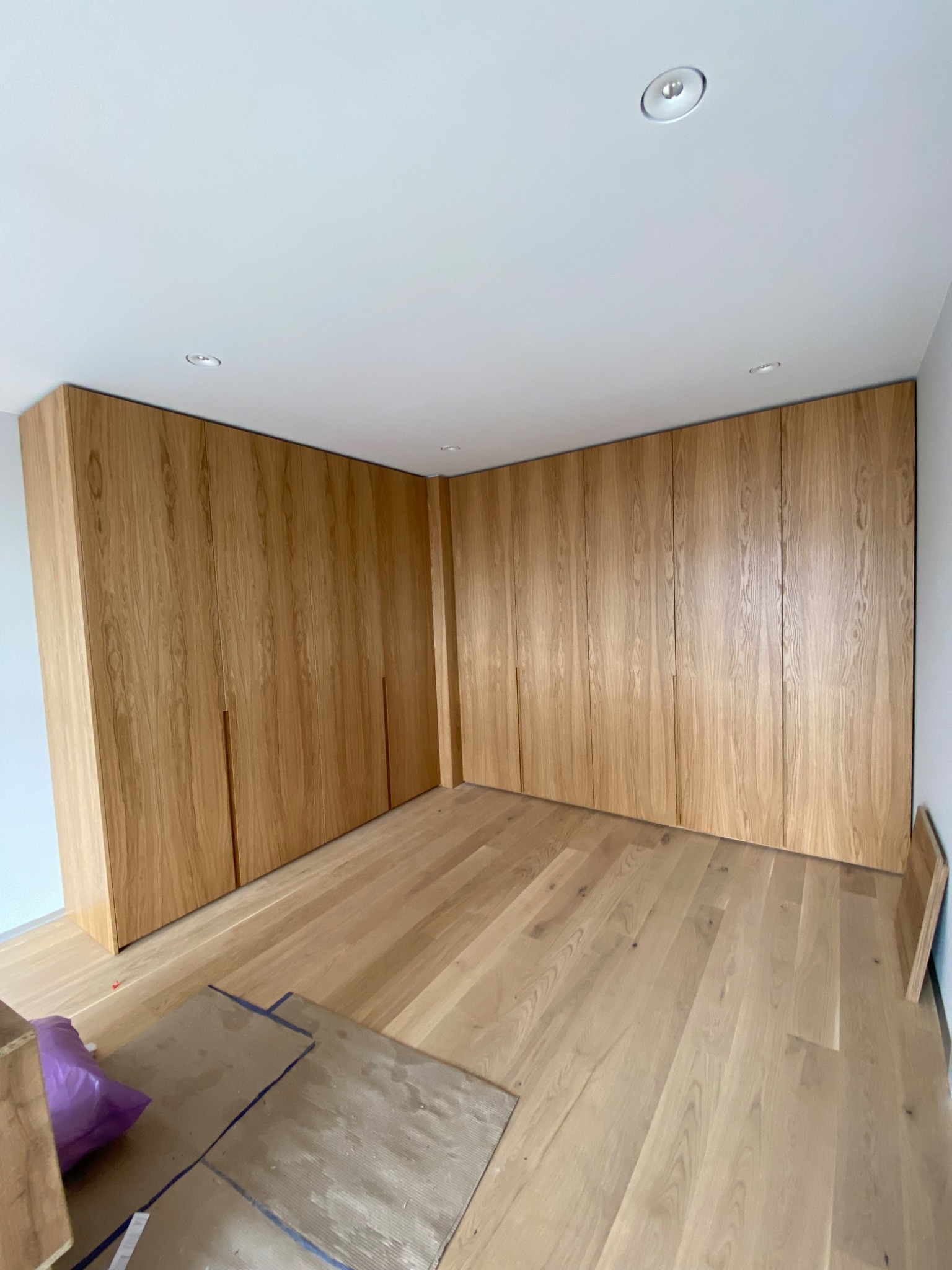 Wnętrze pokoju z zabudową szaf z jasnego drewna, widoczne fragmenty podłogi z paneli i zabezpieczenia podłogi podczas montażu.