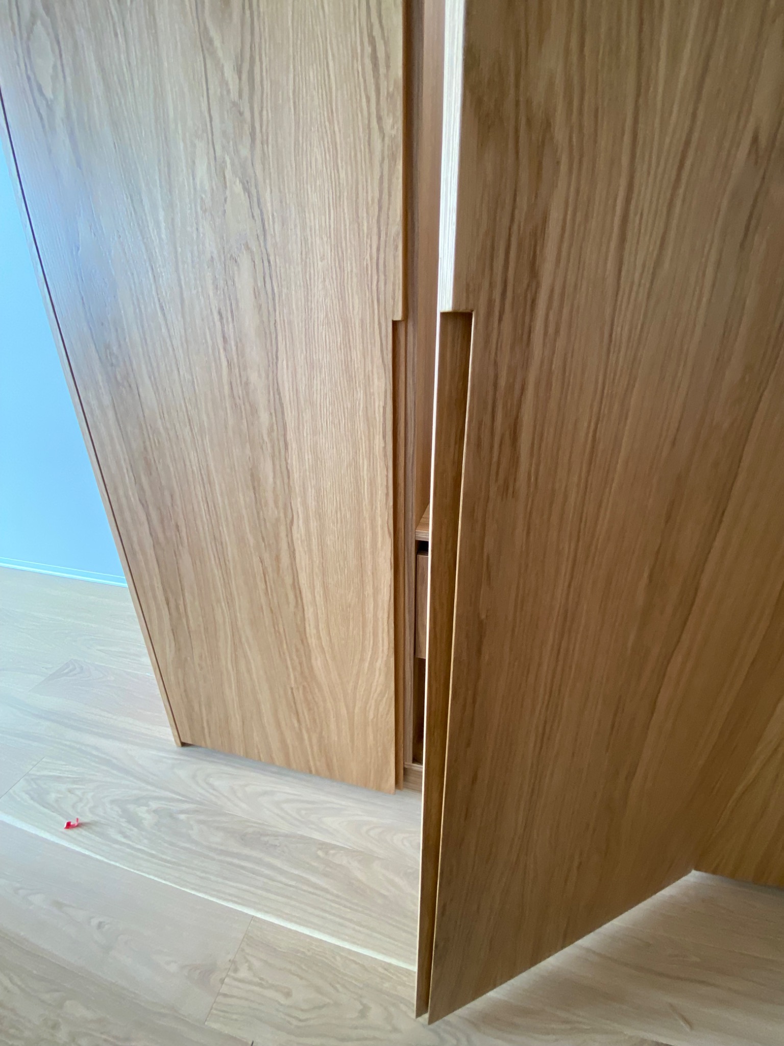 Fragment otwartej szafy z jasnego drewna z ukrytym uchwytem, widoczna podłoga z paneli i fragment niebieskiej ściany w tle.