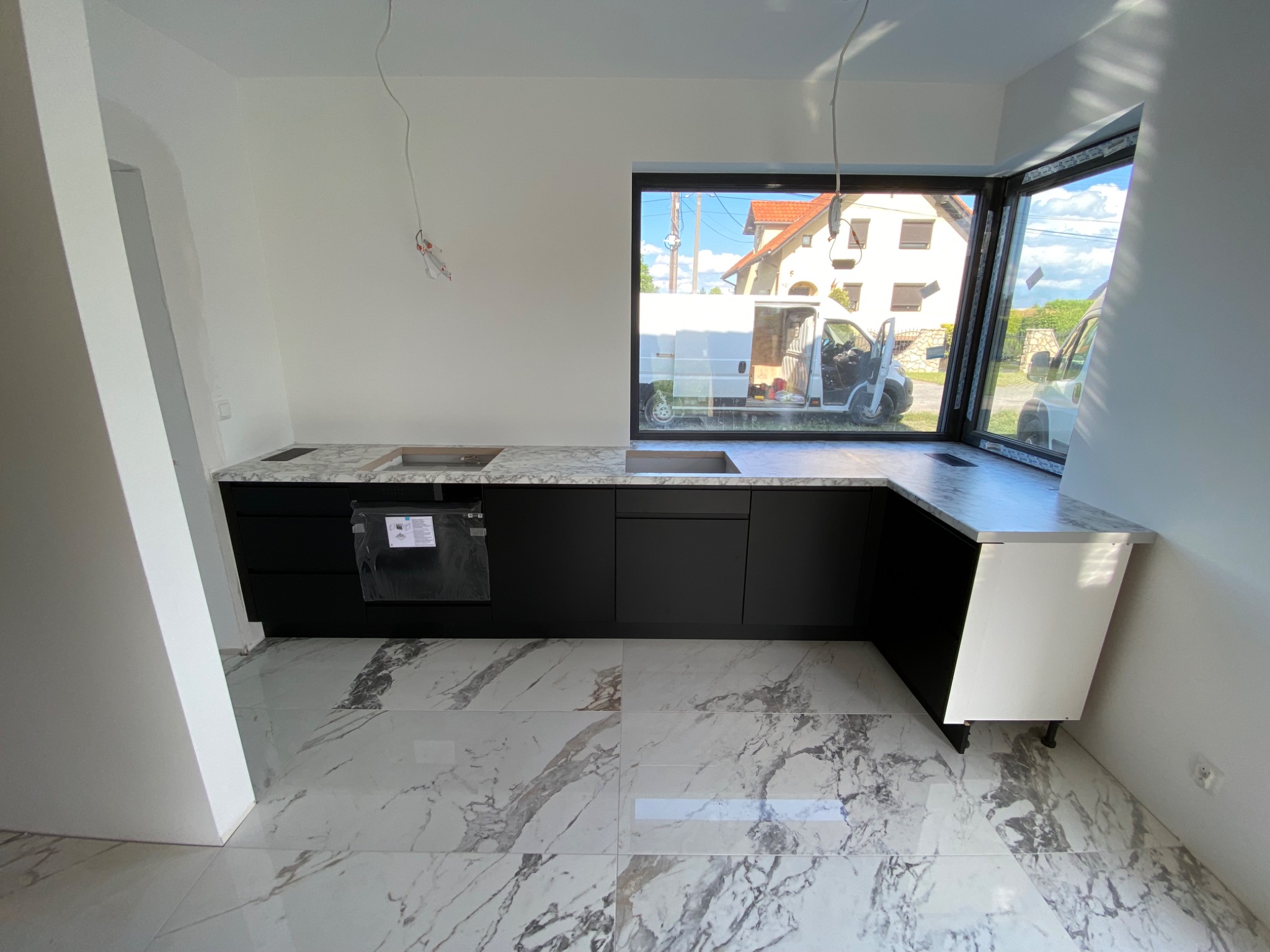 Nowoczesna kuchnia z czarnymi szafkami i marmurowym blatem, narożne okno z widokiem na ulicę, białe płytki podłogowe z szarymi żyłami, przewody zwisające z sufitu.