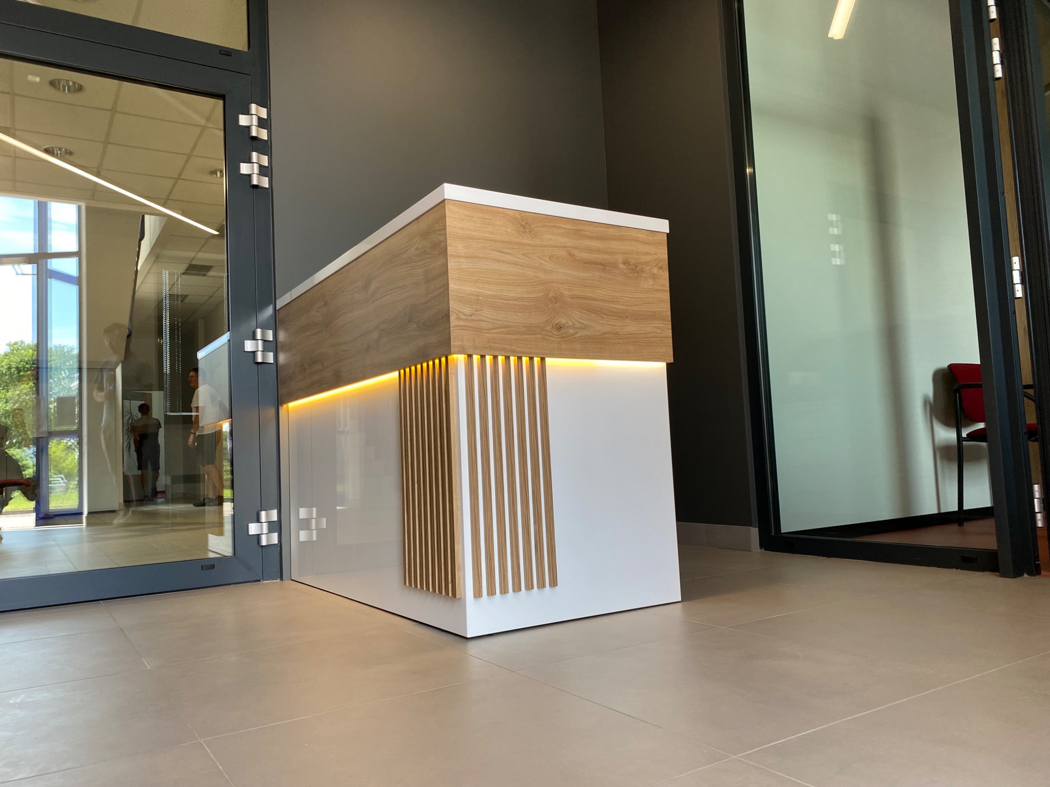 Nowoczesna lada recepcyjna z białymi frontami i blatem z imitacji drewna, podświetlona od dołu, widoczna z perspektywy podłogi w jasnym wnętrzu biurowym z przeszklonymi drzwiami.