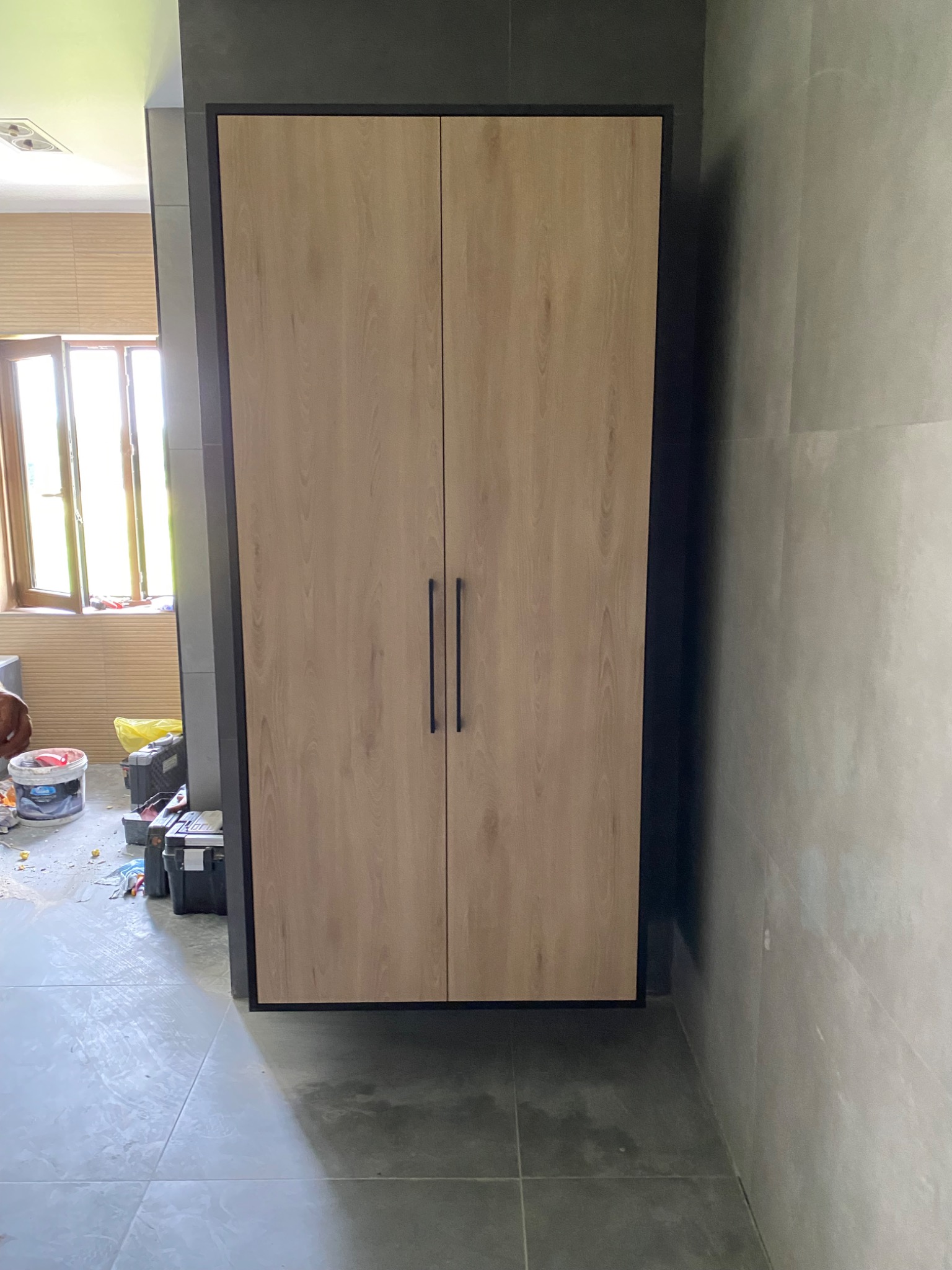 Dwudrzwiowa szafa z jasnego drewna z czarnymi uchwytami, zamontowana na szarej ścianie z płytek, widoczne narzędzia i materiały budowlane w tle.