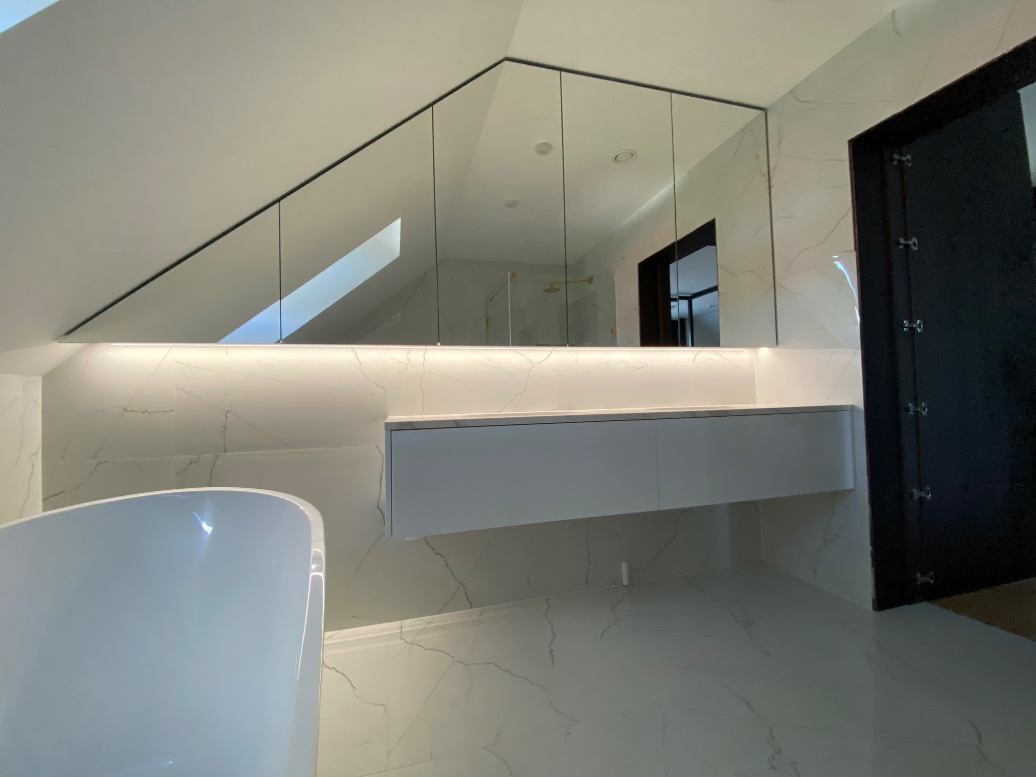 Nowoczesna łazienka na poddaszu z marmurowymi płytkami, lustrami na całą ścianę pod skosem, podświetlaną szafką wiszącą i wolnostojącą białą wanną.