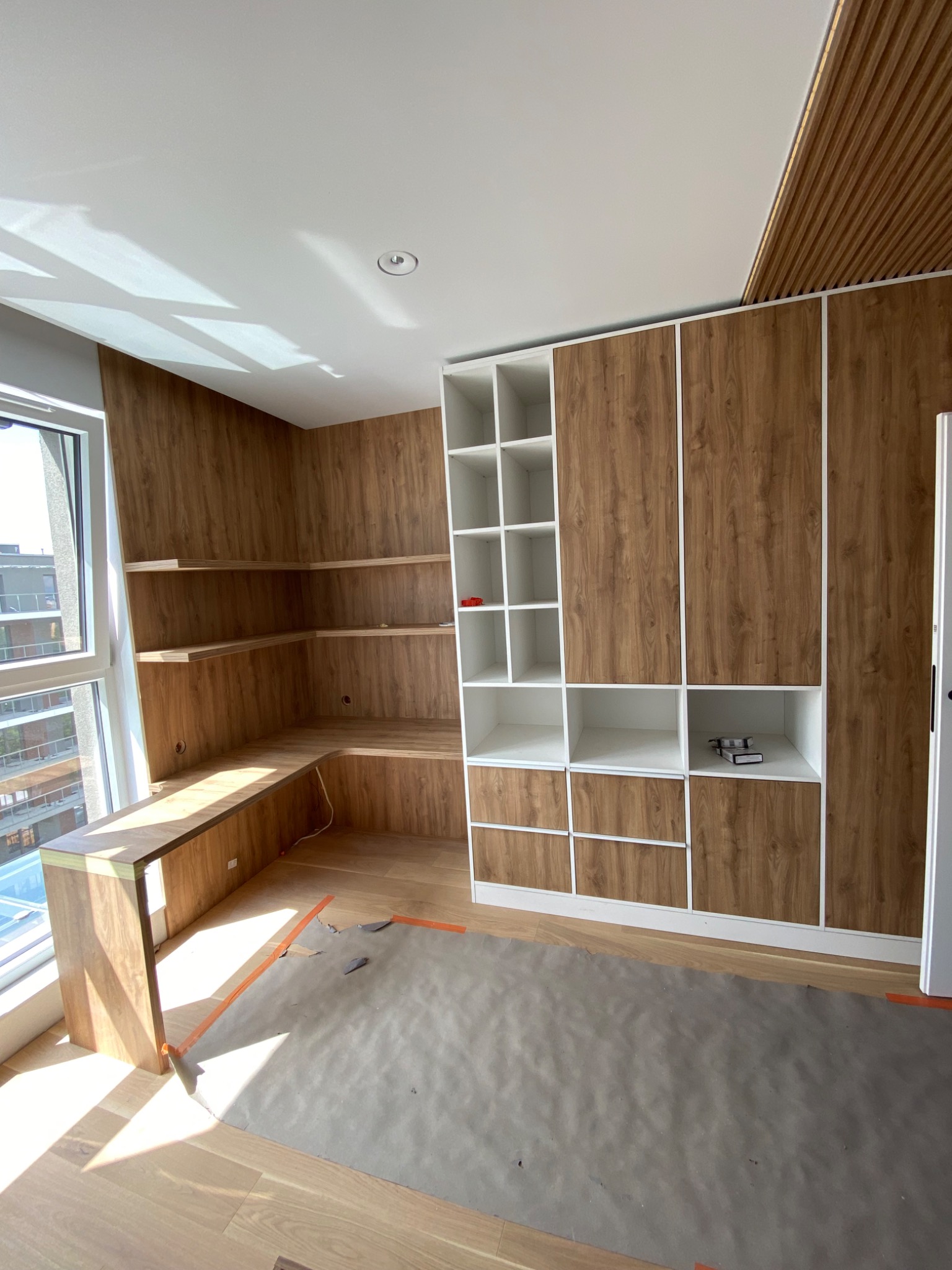 Wnętrze w trakcie wykańczania: zabudowa z półkami i szafkami z forniru drewnopodobnego, widoczny fragment podłogi zabezpieczony folią malarską, światło słoneczne wpadające przez okno.