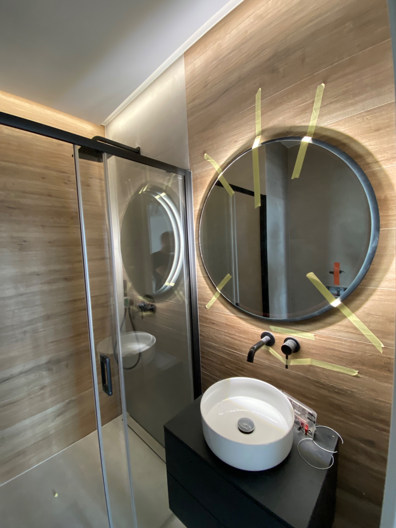 Montaż okrągłego lustra z podświetleniem LED w łazience z drewnopodobnymi płytkami, zabezpieczonego taśmą malarską podczas instalacji baterii umywalkowej nablatowej.