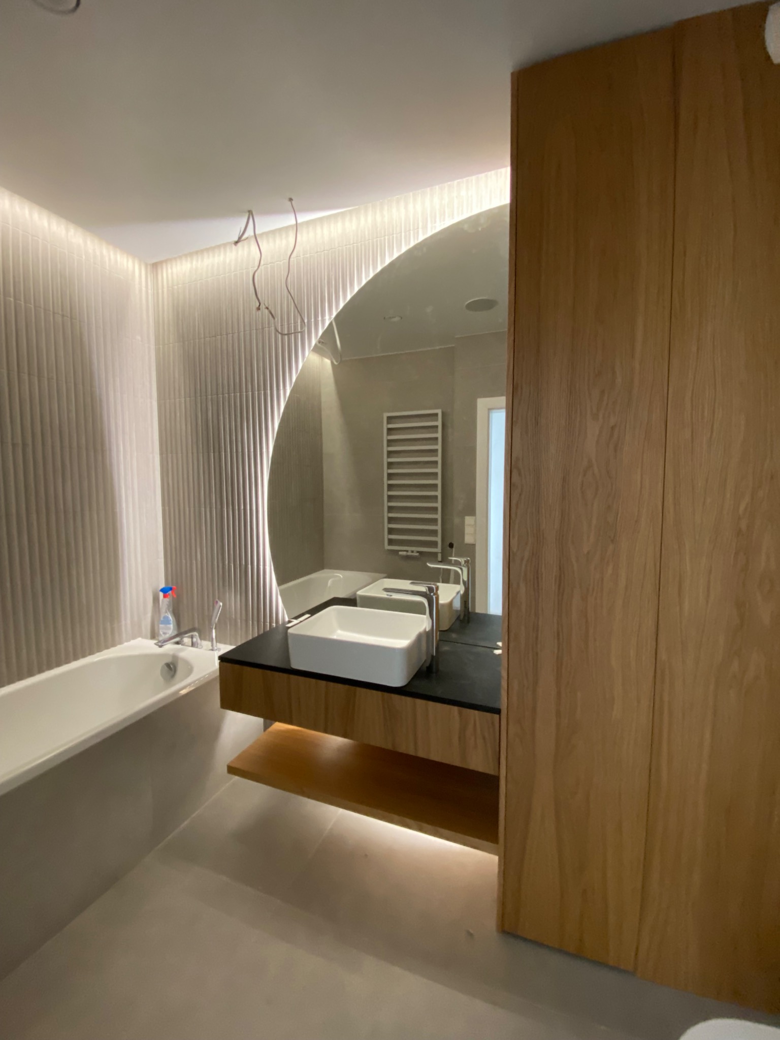 Nowoczesna łazienka z podświetlanym okrągłym lustrem, umywalką nablatową na czarnym blacie i drewnianą zabudową, widoczna wanna i grzejnik drabinkowy w odbiciu, kable wystające z sufitu.