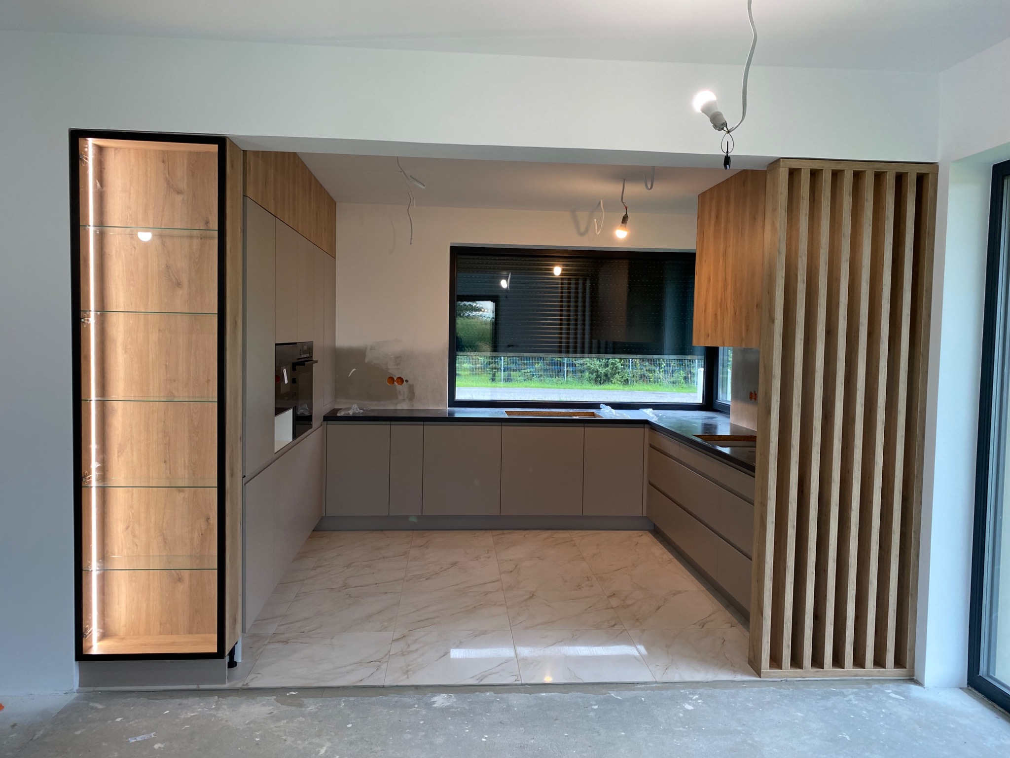 Nowoczesna kuchnia w trakcie montażu w Pszczynie, widoczna zabudowa z oświetloną witryną, drewniane lamele i marmurowe płytki podłogowe, surowy stan wykończenia ścian i sufitu.