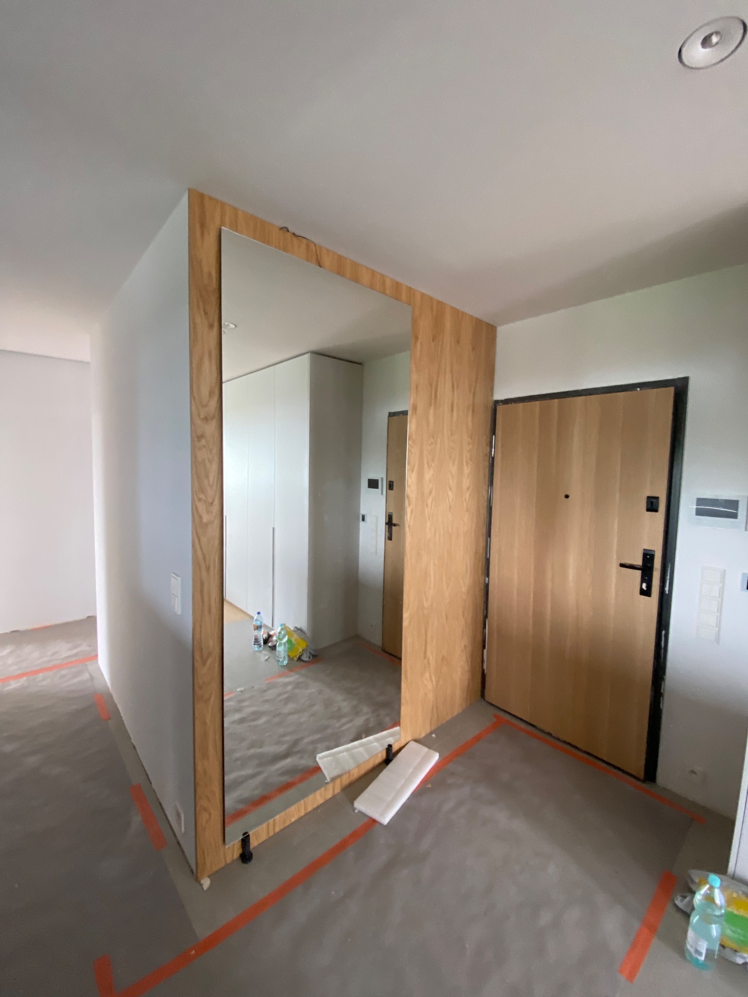 Wnętrze w trakcie wykańczania, widoczna zabudowa z lustrem i drewnianą ramą, drzwi wejściowe z drewnianym fornirem, podłoga zabezpieczona folią i taśmą malarską, w tle biała szafa.
