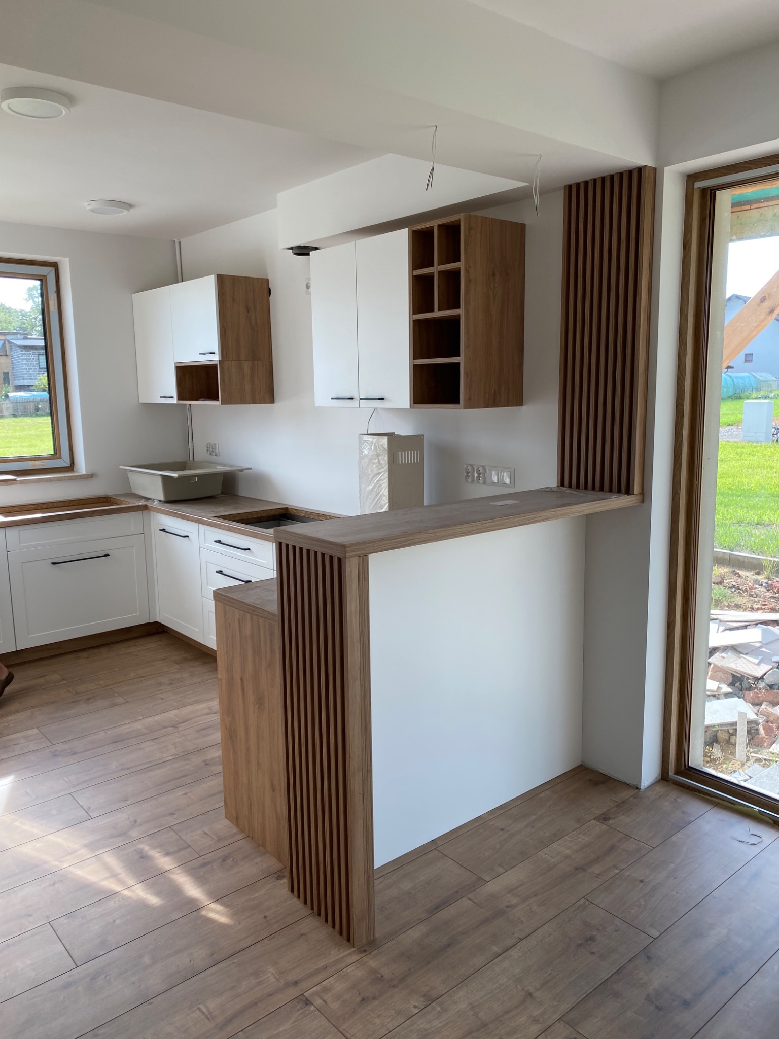 Nowoczesna kuchnia w trakcie montażu, z białymi frontami i drewnianymi akcentami, widoczny barek z lamelami, okno z widokiem na ogród, podłoga wyłożona panelami imitującymi drewno.