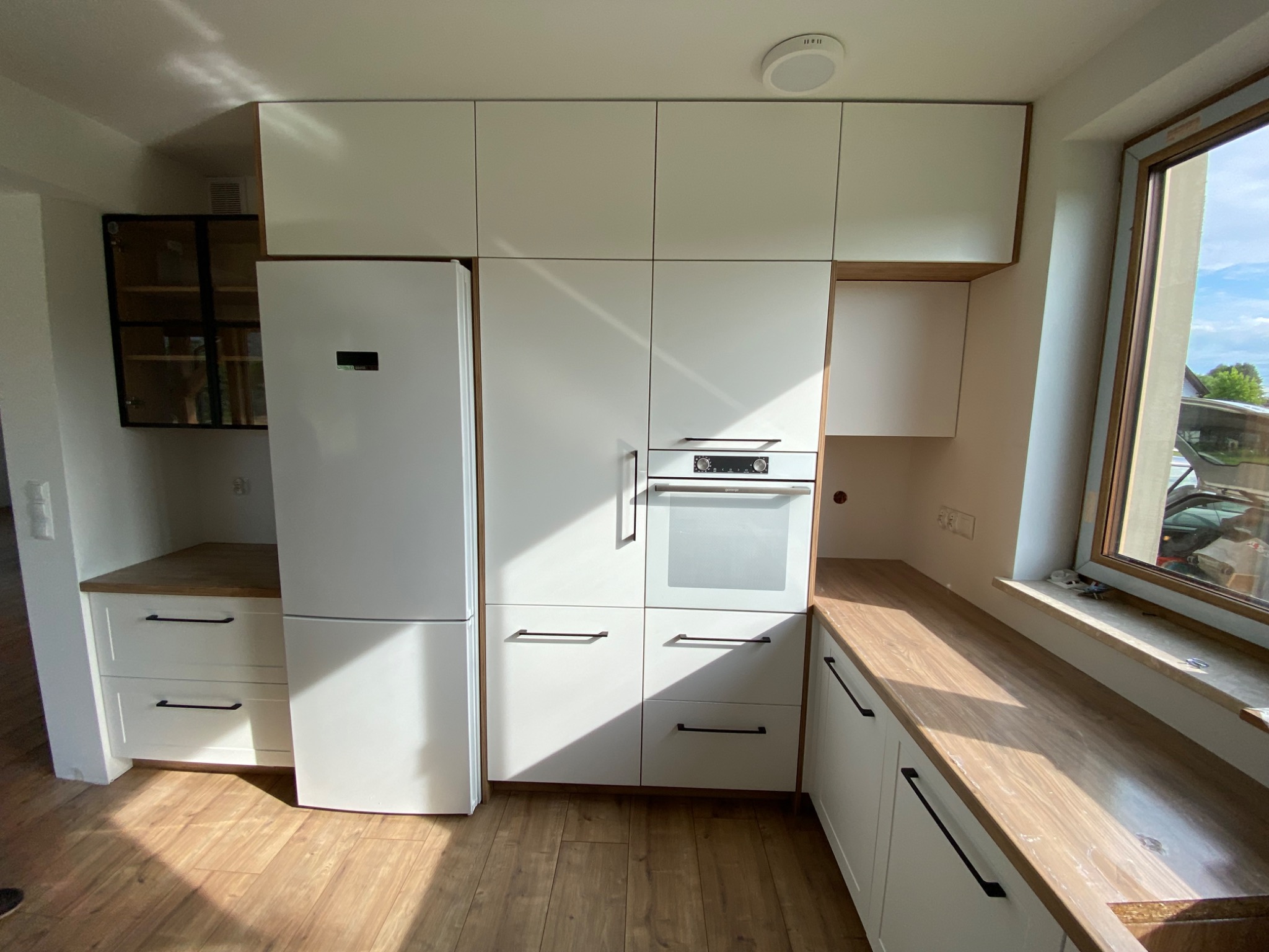 Biała zabudowa kuchenna z drewnianymi blatami, obejmująca lodówkę, piekarnik i szafki wiszące, oświetlona naturalnym światłem wpadającym przez okno.