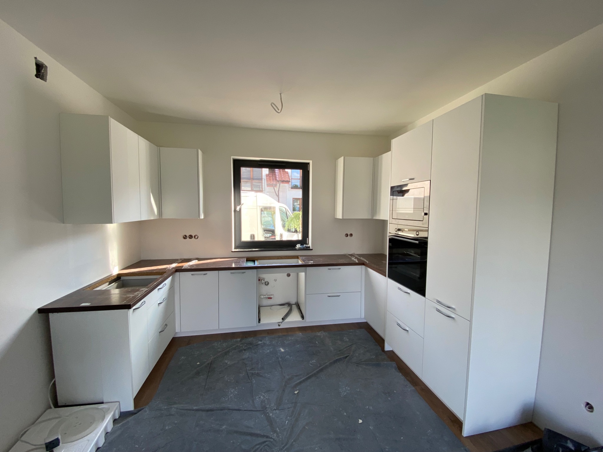 Wnętrze kuchni w trakcie montażu: białe szafki z ciemnym blatem, brakujące elementy instalacji pod zlewem, podłoga zabezpieczona folią, widoczne otwory na gniazdka elektryczne.