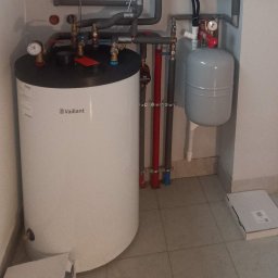 Instal-Grześ Grzegorz Cichy Instalacje wod-kan, gazowe, elektryczne, CO, klimatyzacje, pompy ciepła - Biały zasobnik ciepłej wody Vaillant z szarymi rurami instalacji grzewczej w pomieszczeniu z beżowymi płytkami. Widoczne zawory, manometry i zbiornik wyrównawczy.