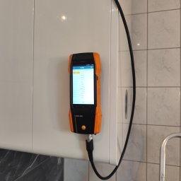 Analizator spalin Testo 300, podłączony przewodem, wyświetla wyniki pomiarów na tle białej szafki i płytek. Urządzenie w obudowie czarno-pomarańczowej.