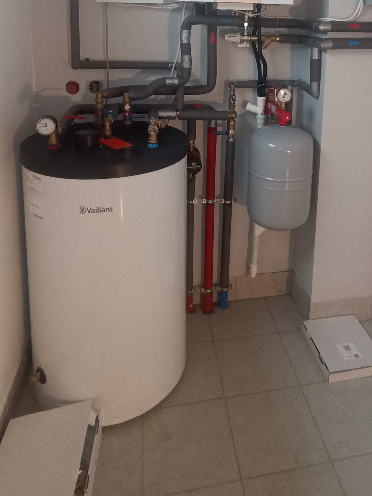Biały zasobnik ciepłej wody Vaillant z szarymi rurami instalacji grzewczej w pomieszczeniu z beżowymi płytkami. Widoczne zawory, manometry i zbiornik wyrównawczy.