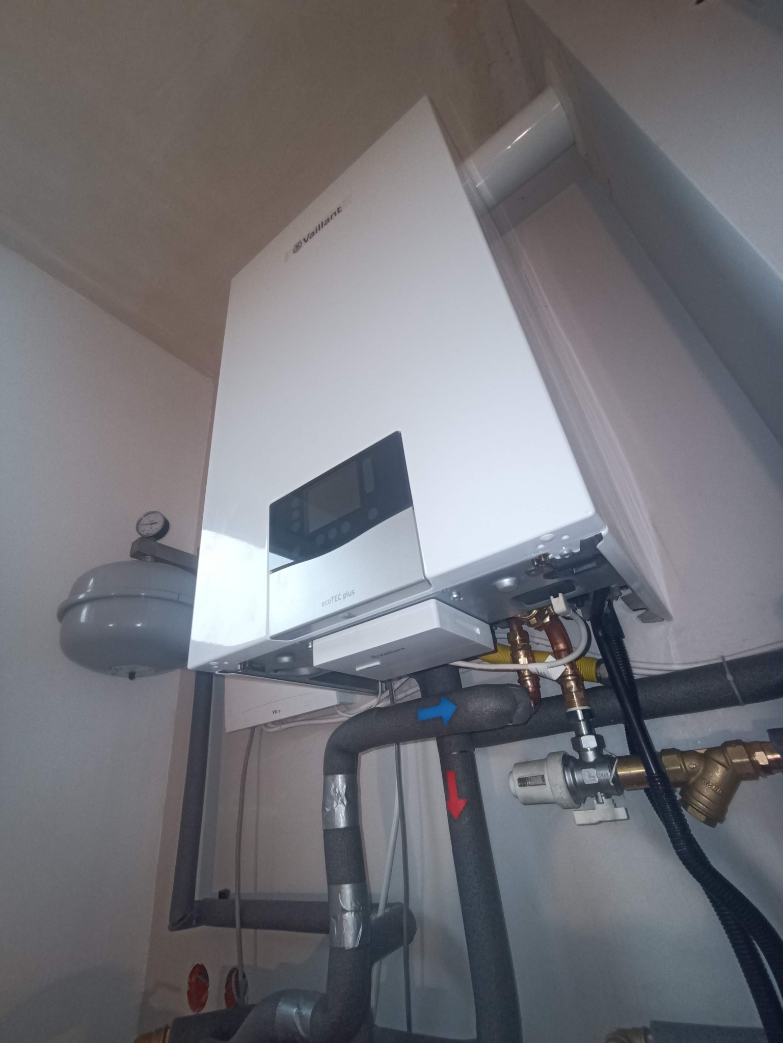 Biały, wiszący piec gazowy Vaillant ecoTEC plus z podłączonymi rurami instalacji grzewczej i gazowej. Widoczne zawory, manometr i izolacja termiczna rur.