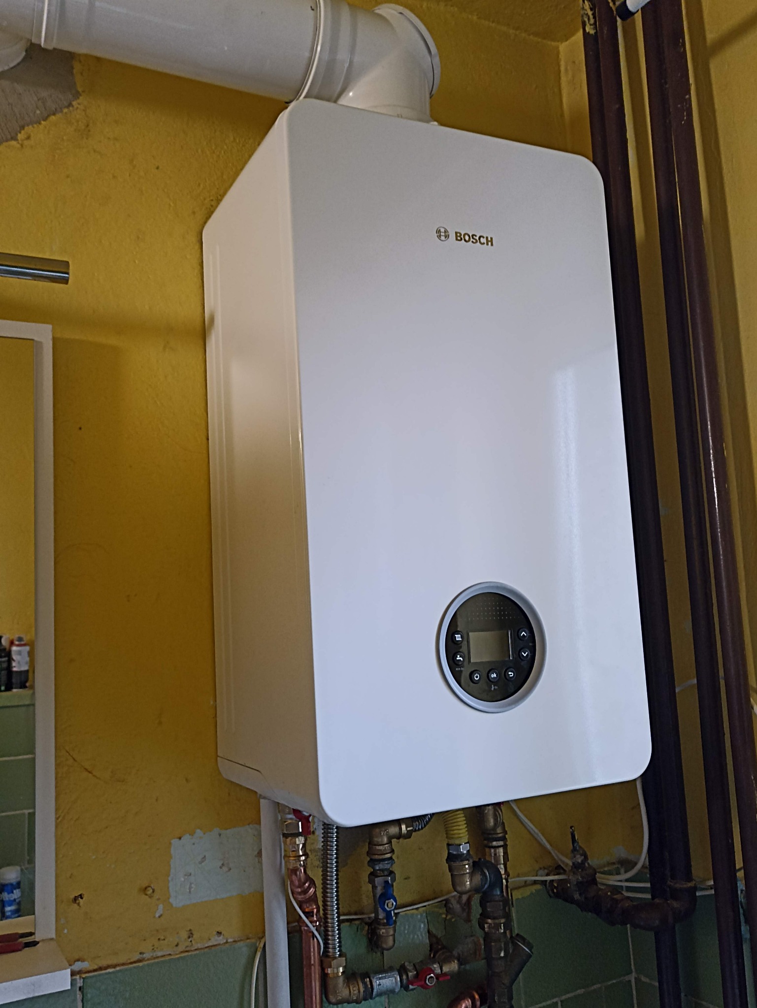 Biały, wiszący piec gazowy marki Bosch z podłączonymi rurami na tle żółtej ściany. Widoczne elementy instalacji i rury odprowadzające spaliny.